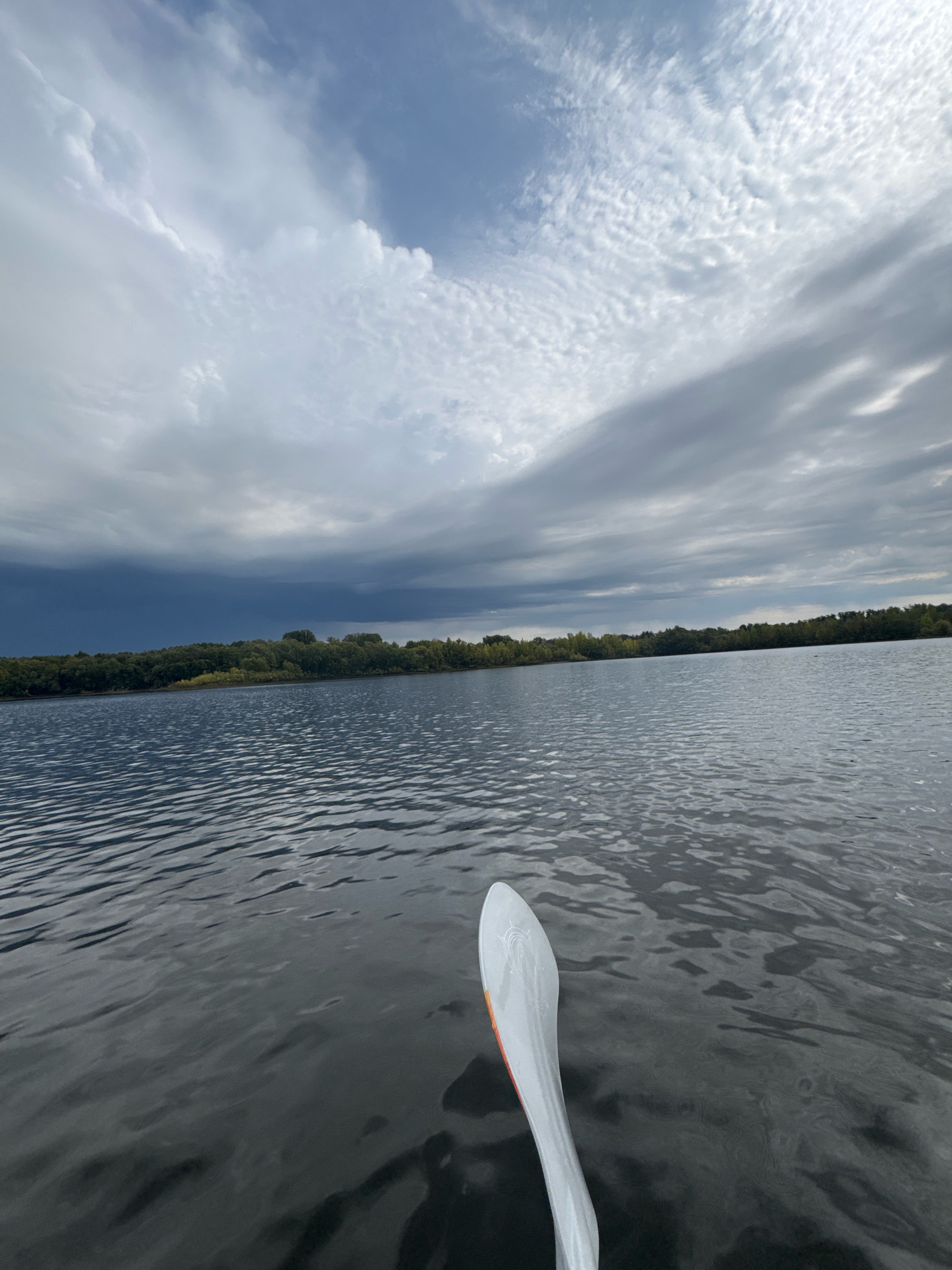 The image depicts a serene lake scene, possibly taken from a kayak or paddleboard, given the presence of a paddle in the foreground. The water's surface is calm with gentle ripples, reflecting the overcast sky. The distant shoreline is lined with a dense forest of green trees, suggesting a natural or park-like setting. The sky is dramatic, with a mix of heavy, dark clouds in the distance, hinting at potential rain, and lighter, wispy clouds overhead. This could be a scene from a lake in or around Ufa, Russia, given the general geography and climate. There are no people visible in the frame other than potentially the person taking the photo.