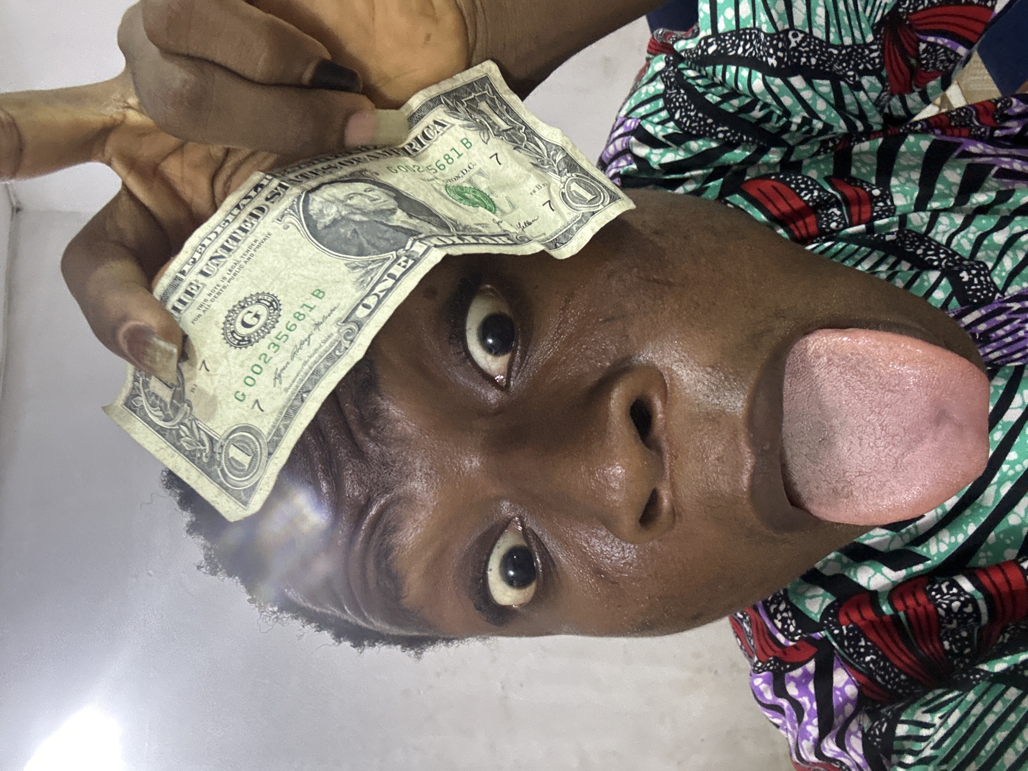 A close-up, rotated image captures a person's face, displaying a playful and wide-eyed expression with their tongue sticking out to the side. A dark-skinned hand holds a crumpled US one-dollar bill over the person's forehead, partially obscuring one eye. The dollar bill, a Series 2017 note with the serial number G00235681 B, features the inscription "THIS NOTE IS LEGAL TENDER FOR ALL DEBTS, PUBLIC AND PRIVATE" and "THE UNITED STATES OF AMERICA." The person is wearing a vibrant, multi-patterned garment, possibly made of African print fabric, characterized by geometric shapes and stripes in shades of green, black, white, red, and purple. The background is a plain, light-colored surface, suggesting an indoor setting with artificial lighting, as indicated by a slight glare on the lower left. The overall mood conveyed is humorous and informal.