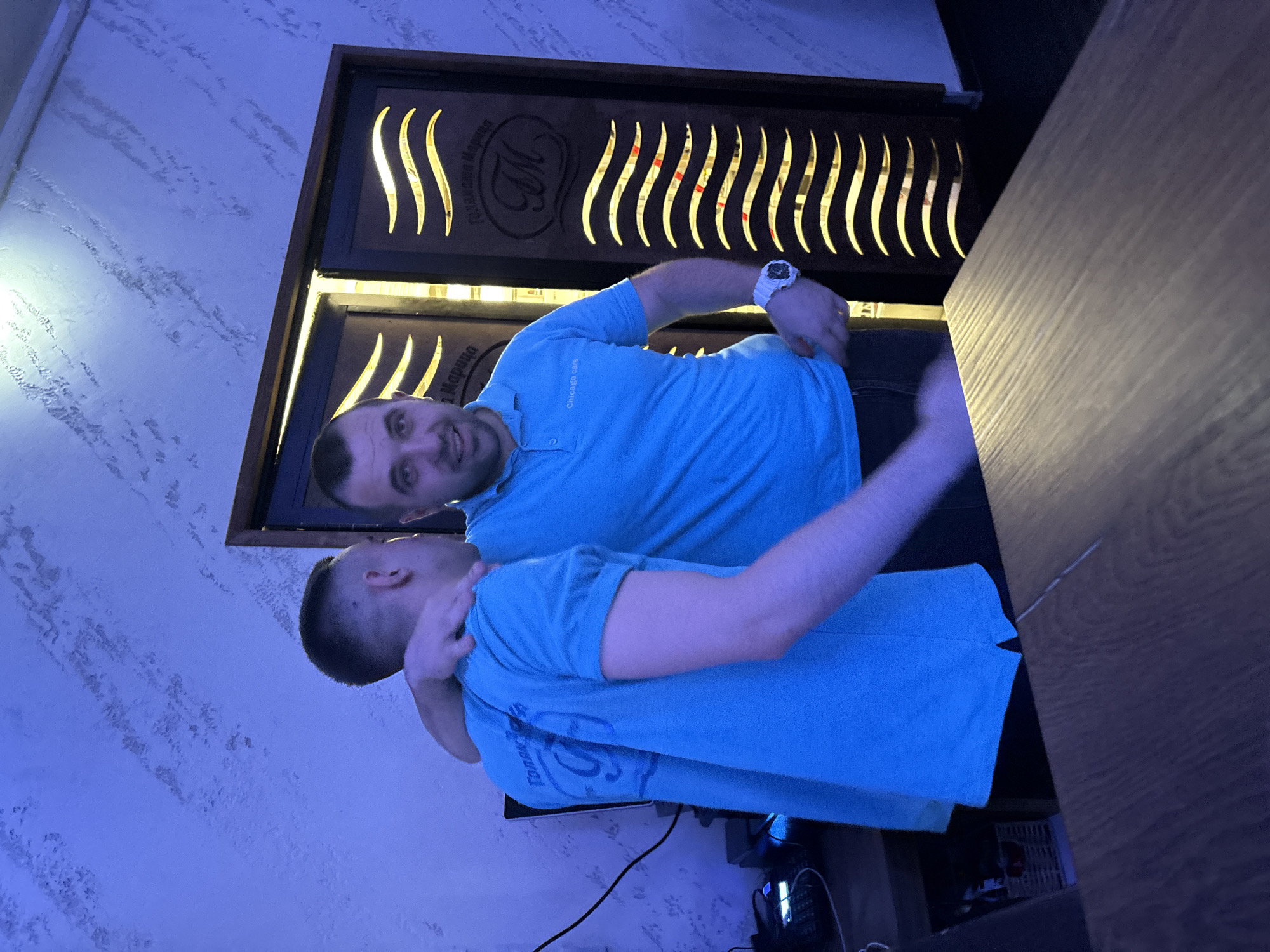 Two men, both wearing blue polo shirts, are embracing. One man has his arm around the other's neck, looking at the camera with a smile. The other man has a buzz cut and a blank expression. They are standing inside, illuminated by blue lighting. Behind them is a wooden structure with decorative cutouts that resemble stylized waves and the Cyrillic letter "M." The structure also features the words "Тракийски Марципан" (Trakiiski Martzipan) and "Марципан" (Martzipan). A portion of a dark wooden table is visible on the right side of the frame. Cables and electronic devices are visible on the lower edge of the frame, suggesting a technological setup. The location is Plovdiv, Bulgaria, based on the inscription "Тракийски Марципан" which is the name of a store located in the city.
