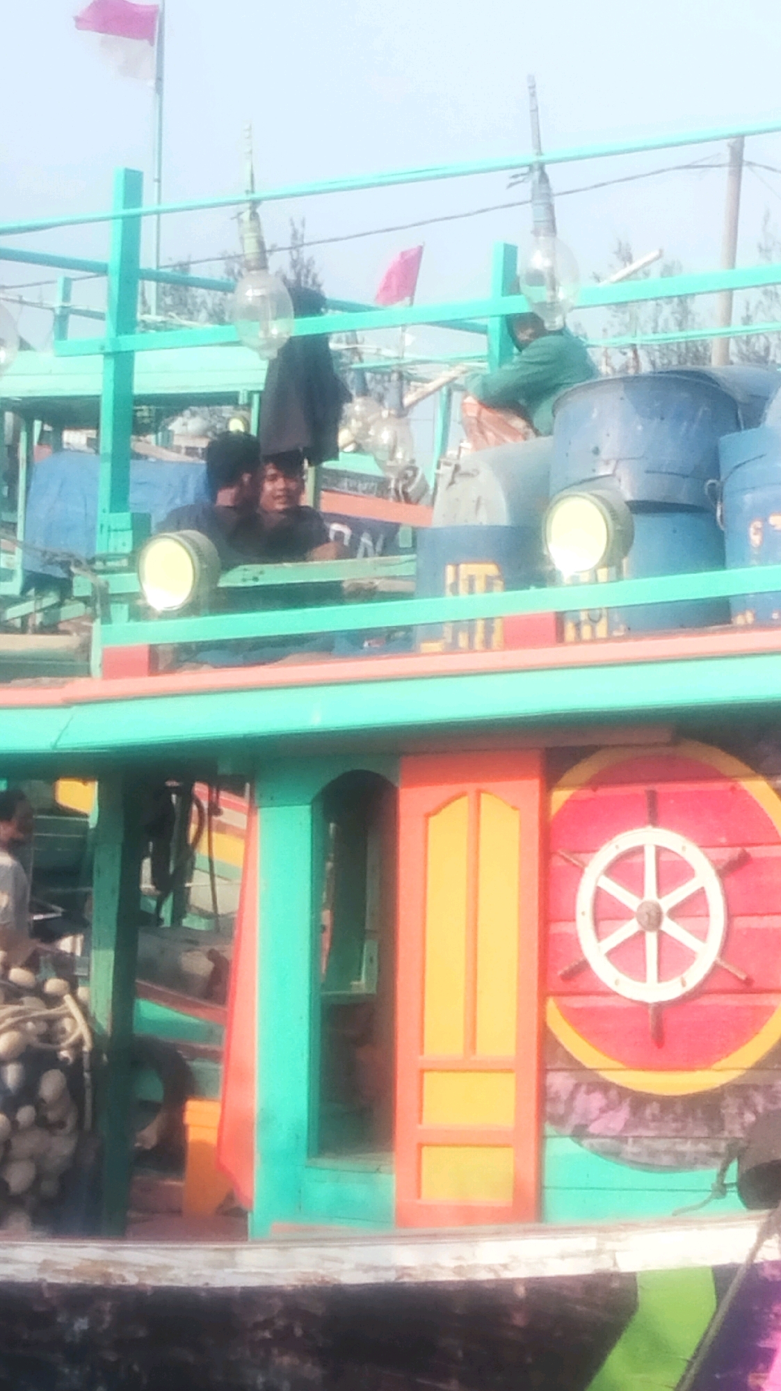 In Palang, Indonesia, a fishing boat with vibrant turquoise, red, and yellow paint is docked. On the boat's deck, several individuals are present. Two men, wearing dark shirts, are looking towards the camera. Another person, wearing a green jacket, is seated on the boat. Large, clear glass light bulbs are suspended from a turquoise frame. The boat features a decorative ship's wheel painted on its side. The background shows a hazy sky with a few trees and flags.