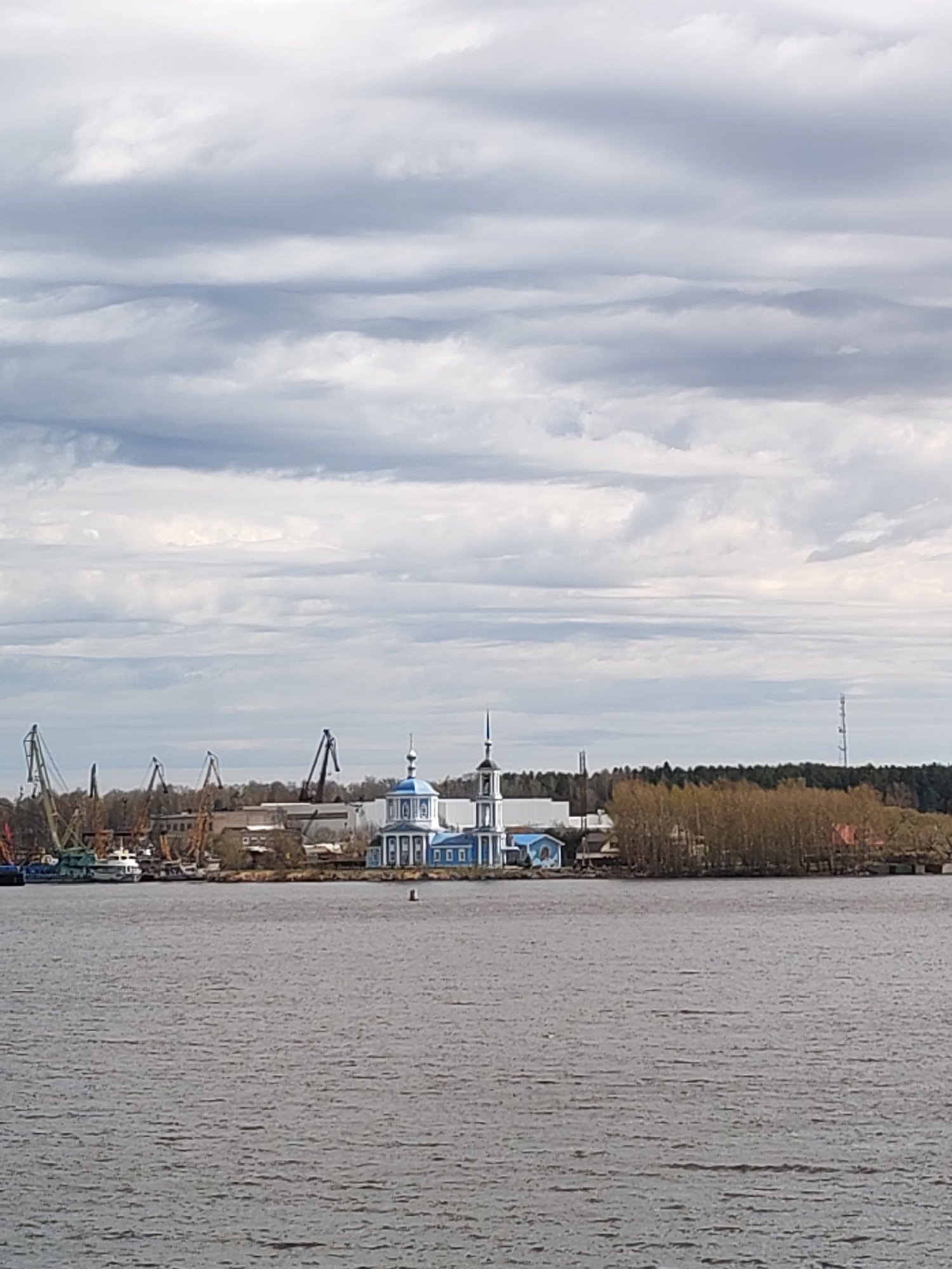 The image shows a body of water with a shoreline in the background. On the shoreline is a blue and white building, likely a church, with a dome and a spire. Adjacent to the church are industrial cranes and buildings. Beyond that is a line of trees and a communications tower. Above, the sky is filled with layered grey and white clouds. Burtsevo, Russia is a possible location given the presence of the Volga River, Russian-Orthodox Church structures, and water crane infrastructure in the area.