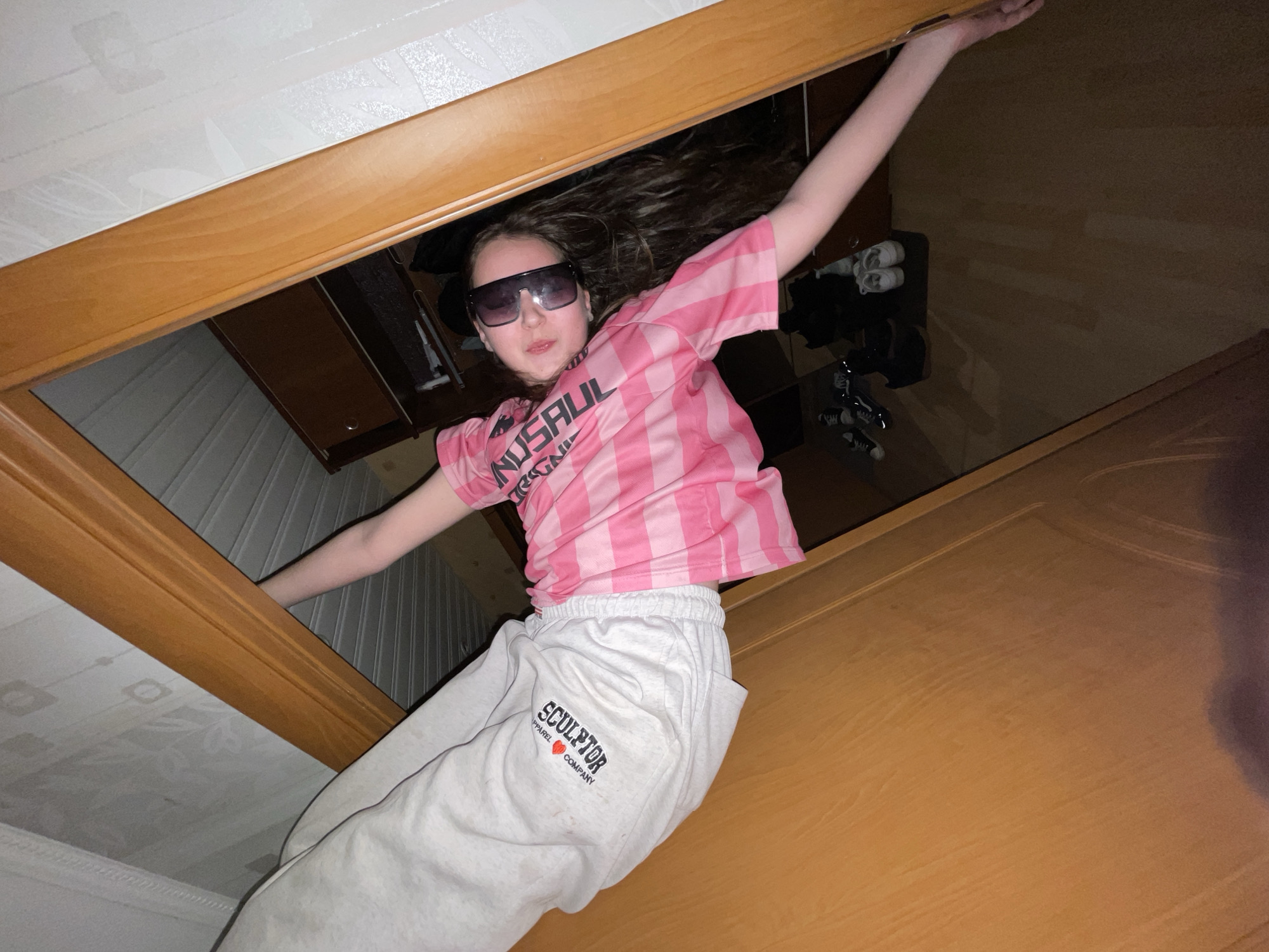 The image captures a young person, likely a child or teenager, from a low-angle perspective looking upwards through a wooden frame. The subject is centrally positioned within this frame, which appears to be part of a doorway or an opening in a room, perhaps a loft or an unusual architectural feature.
The person is wearing large, dark rectangular sunglasses, a pink and white striped short-sleeved t-shirt, and light gray sweatpants. Their arms are outstretched, with the right arm extending upwards to grasp the wooden frame, and the left arm extended outwards to the side. Long, dark hair frames their face. The pose suggests they are either hanging from, climbing through, or playfully leaning within the opening, creating an illusion of being suspended.
The visible text on their clothing includes "UNUSUAL" (inverted and partially visible) in white on the pink striped t-shirt. On the gray sweatpants, the brand "SCULPTOR APPAREL COMPANY" is clearly printed in dark letters with a small heart logo beneath it.
The background above the wooden frame shows a white ceiling with a subtle embossed or textured pattern. Below the subject, a dark, reflective surface, likely a floor or furniture, reflects dim light and several pairs of shoes, including white sneakers and dark footwear. A light brown wooden door or wall panel is visible on the right side of the frame. The indoor setting is dimly lit, suggesting artificial light or evening hours. The overall mood is playful and energetic, captured with a unique photographic angle in Novoe Devyatkino, Russia.