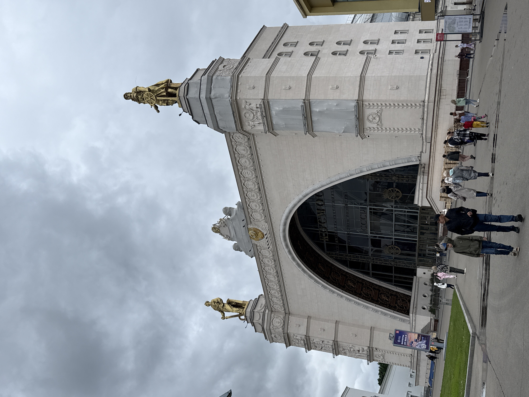 The image shows the Pavilion "Cosmos" at the All-Russian Exhibition Center (VDNKh) in Moscow, Russia. The pavilion is a large, light-colored building with a grand arched entrance and two golden statues on top. The sky is overcast with grey clouds. In front of the pavilion, a group of people are walking on a paved area. Some are walking towards the camera, while others are walking away. There is some greenery on the left side of the image, and a sign with Russian text is visible on the left. The overall scene depicts a public space with a notable architectural landmark and a scattering of visitors.