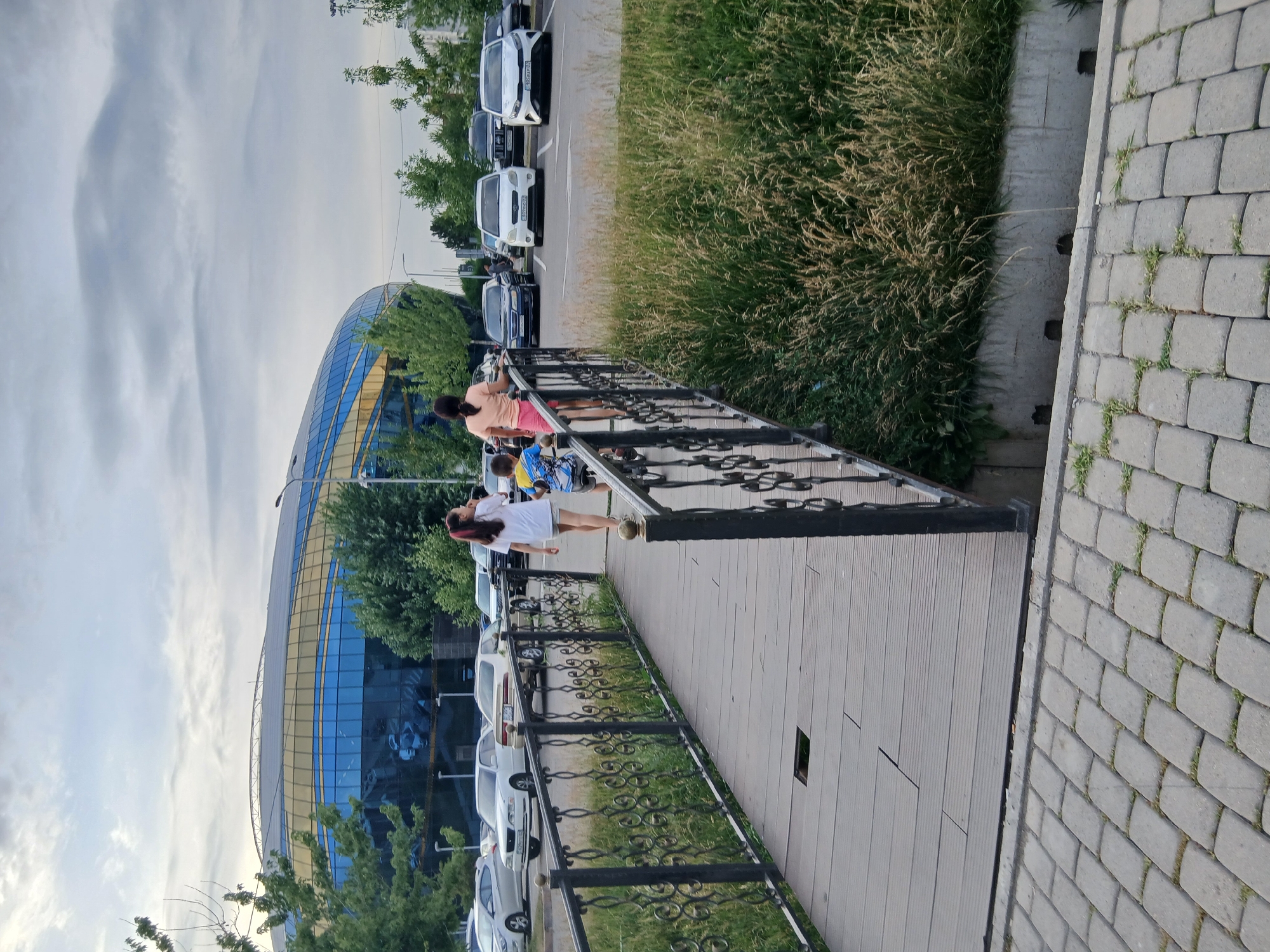 The image depicts a scene with people, cars, and a modern building. In the foreground, a wooden walkway with a black metal railing is visible. Several people are walking on the walkway, including a woman in a white top and shorts, and another woman in a peach-colored top and pink shorts. Further down the walkway, a child is visible with a backpack.
Behind the walkway, a row of cars is parked. The cars are of various colors, including white, black, and blue. Beyond the cars, a large, modern building with a curved, glass facade is prominent. The building's windows are blue, and it appears to have a yellow trim. Green trees surround the building, and a grassy area is also visible.
To the right of the walkway, a paved area with grey bricks is present, with some grass growing between the stones. The sky is overcast with light clouds. The overall impression is of an outdoor urban setting, possibly a public space or commercial area, during daylight hours. No specific text is visible in the image. The scene appears to be tranquil, with people engaged in casual movement.