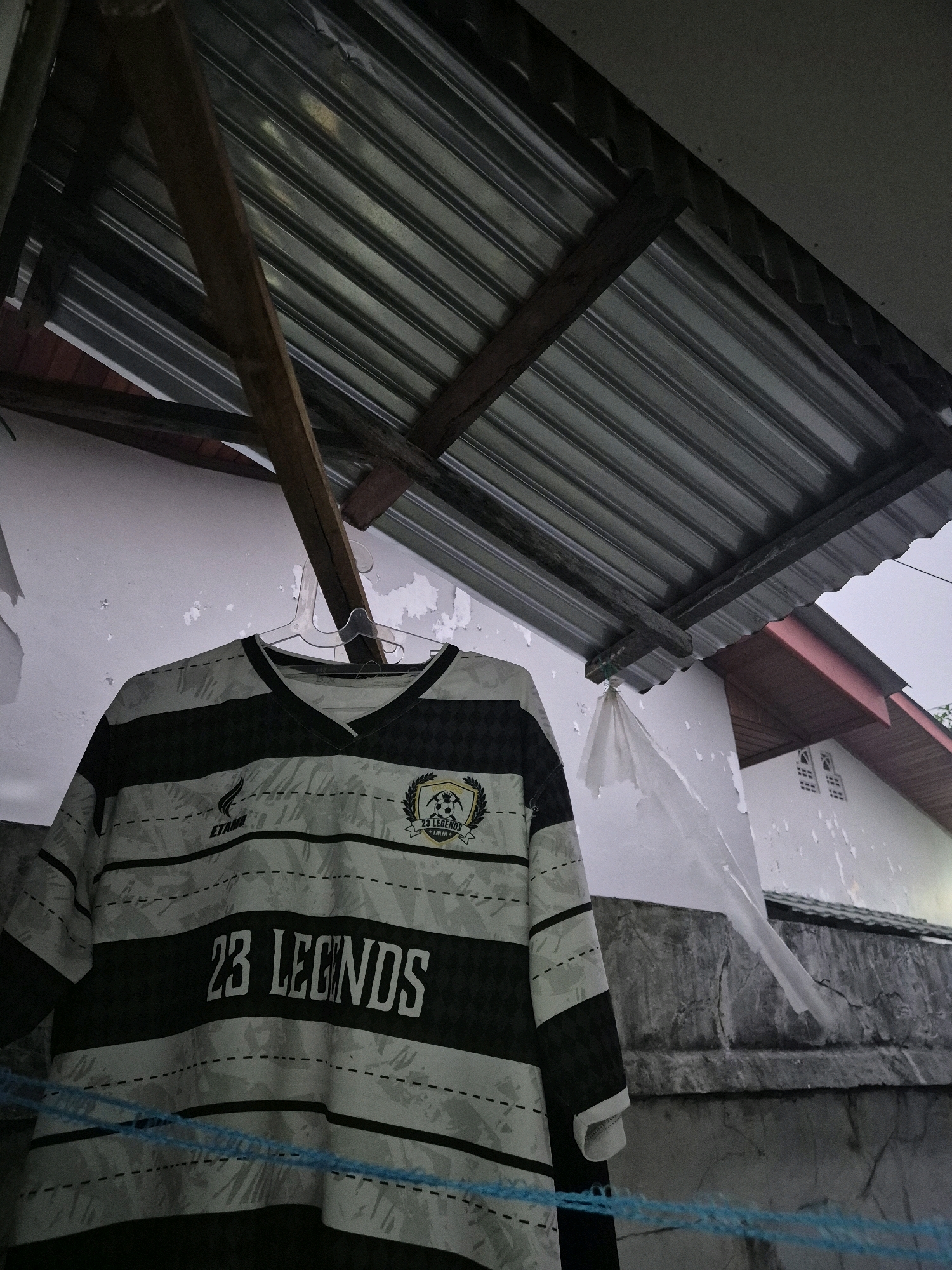 A soccer jersey with black and white horizontal stripes hangs on a hanger. The jersey has the text "23 LEGENDS" in white lettering across the chest. A logo with a soccer ball and the words "23 LEGENDS" is visible on the right side of the jersey. The jersey is outdoors, possibly drying, under a corrugated metal roof supported by wooden beams. A white wall with peeling paint is visible behind the jersey, and a concrete wall is in the background. The scene appears to be in an outdoor residential area in Bontang, Indonesia.