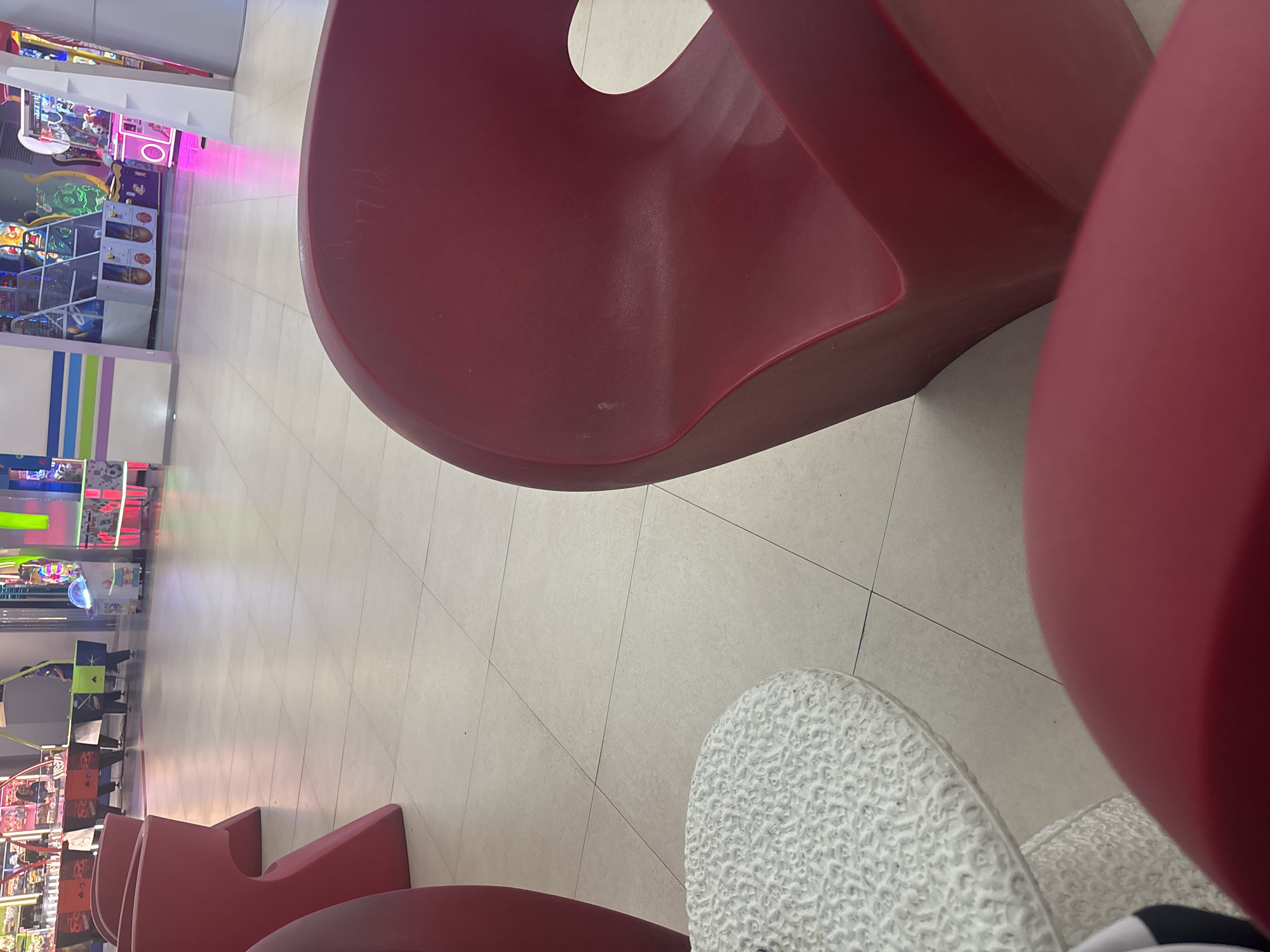 The image, rotated 90 degrees counter-clockwise, depicts an indoor public space, likely an arcade or entertainment center, viewed from a low, abstract perspective. In the immediate foreground and midground, several large, sculptural red chairs or benches with organic, curved shapes dominate the frame. The smooth, matte surface of these chairs shows a few minor scuffs and, on one chair, some faint, indistinct markings or etched text that is difficult to decipher. A white, textured object, possibly another seat or a footrest, is partially visible in the bottom right corner. The floor throughout the area is covered with light-colored square tiles.
In the background, a vibrant arcade setting unfolds with numerous game machines. Bright, artificial lights in shades of pink, green, and blue illuminate various gaming cabinets. Visible among them are at least two basketball shooting games. The walls feature sections with colorful horizontal stripes (white, light blue, purple, light green). While no people are clearly visible, the arrangement of seating and entertainment machines indicates a venue designed for leisure and play. The setting suggests an energetic atmosphere typical of an indoor amusement facility, likely located in Yerevan, Armenia. The artificial lighting makes the time of day indeterminate.