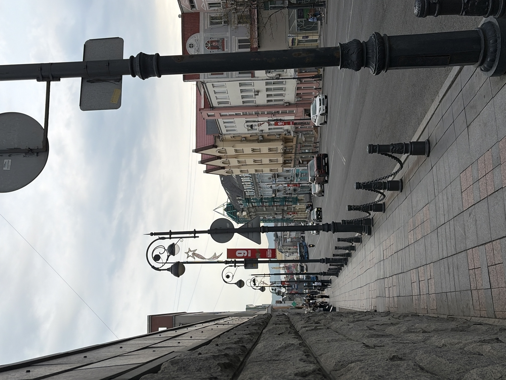 This photo shows a street scene in Vladivostok, Russia. It features a row of decorative, black lampposts along a sidewalk made of large gray slabs. The lampposts have chained barriers. A gray stone wall runs along the bottom of the image. Several cars are visible in the street, with a few people walking on the sidewalk. Banners and signs are displayed on some lampposts, including a red banner with the date "9 MAЯ" (May 9th), likely referring to Victory Day celebrations. Buildings of various heights and colors line the opposite side of the street. The sky is overcast and gray.