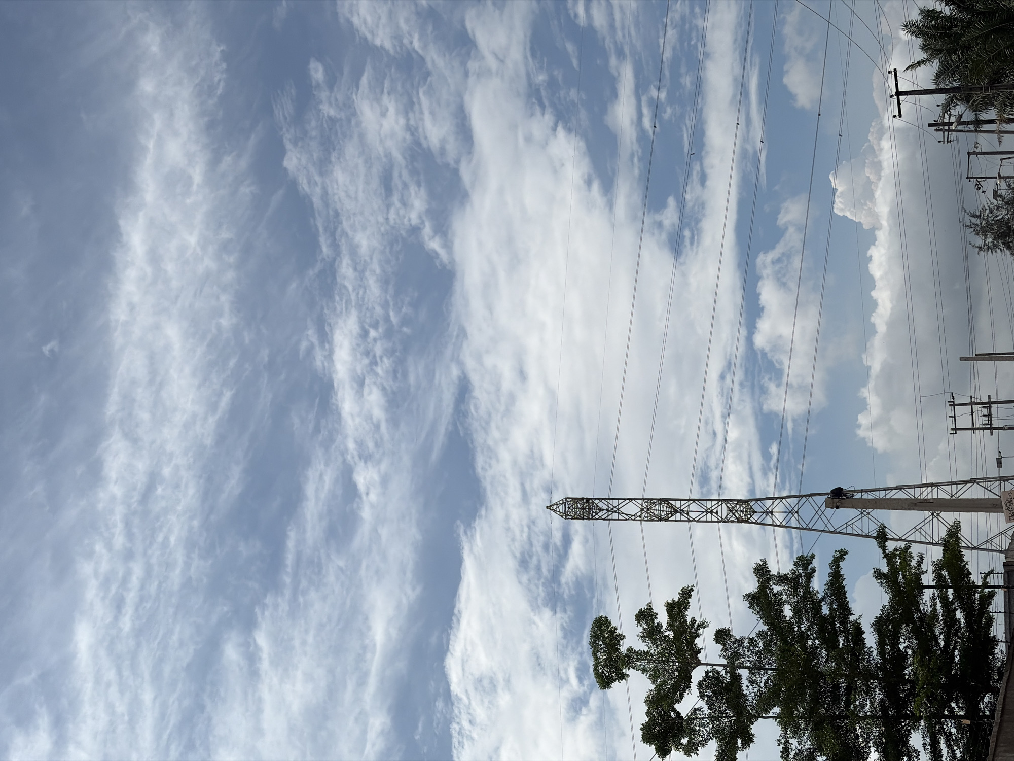 Igboefon, Nigeria: Low angle view of a cloudy sky with cirrus formations, interspersed with patches of blue. Overhead electrical transmission towers and lines are visible. Trees are present in the foreground. Utility poles line the right side of the frame.