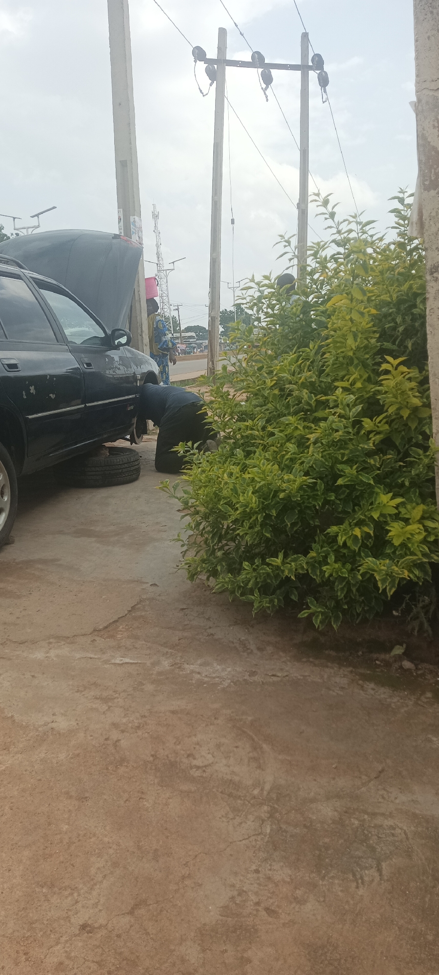 In Bukuru, Nigeria, a dark-colored sedan is parked with its hood open and a tire removed, suggesting mechanical work is being performed. A person in dark clothing is kneeling beside the car, seemingly engaged in repairing it. Another individual, wearing a blue and yellow patterned garment, stands in the background, observing the process. Behind them, utility poles with electrical wires stretch across the cloudy sky. Lush green foliage partially obscures the scene on the right side, hinting at an outdoor, possibly roadside, location. The ground appears to be paved with concrete.