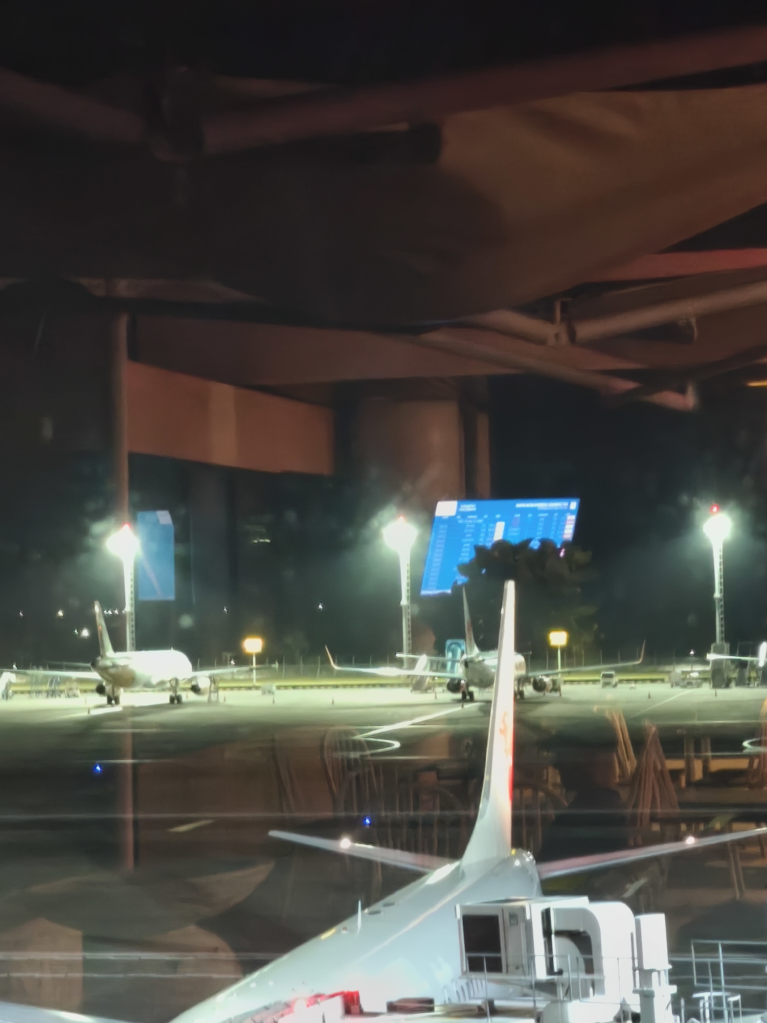 At night, at an airport in Kuta, Indonesia, several airplanes are parked on the tarmac. A large digital display board shows flight information in blue and white. The scene is illuminated by runway lights and the overhead lighting of the airport terminal. The image is taken from inside the terminal, looking out through a window, which reflects some of the interior lighting.