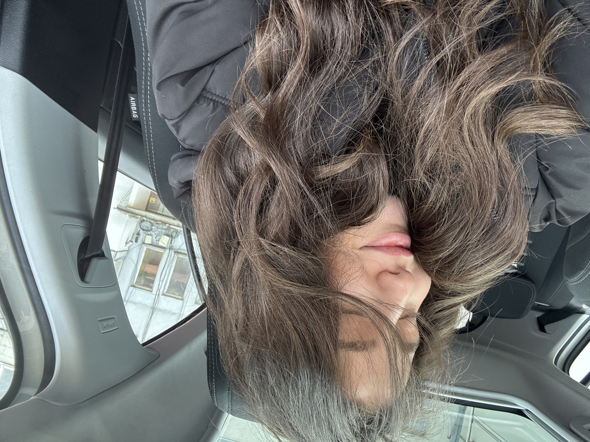 The image is taken from inside a vehicle, showing a person with long brown hair leaning backward. The person is wearing a black jacket. The interior of the car is visible, including the headrest, dashboard, and the passenger-side A-pillar with an "AIRBAG" label. Outside the car window, a building with windows can be seen. The location in the city of Unknown, Unknown is unknown.