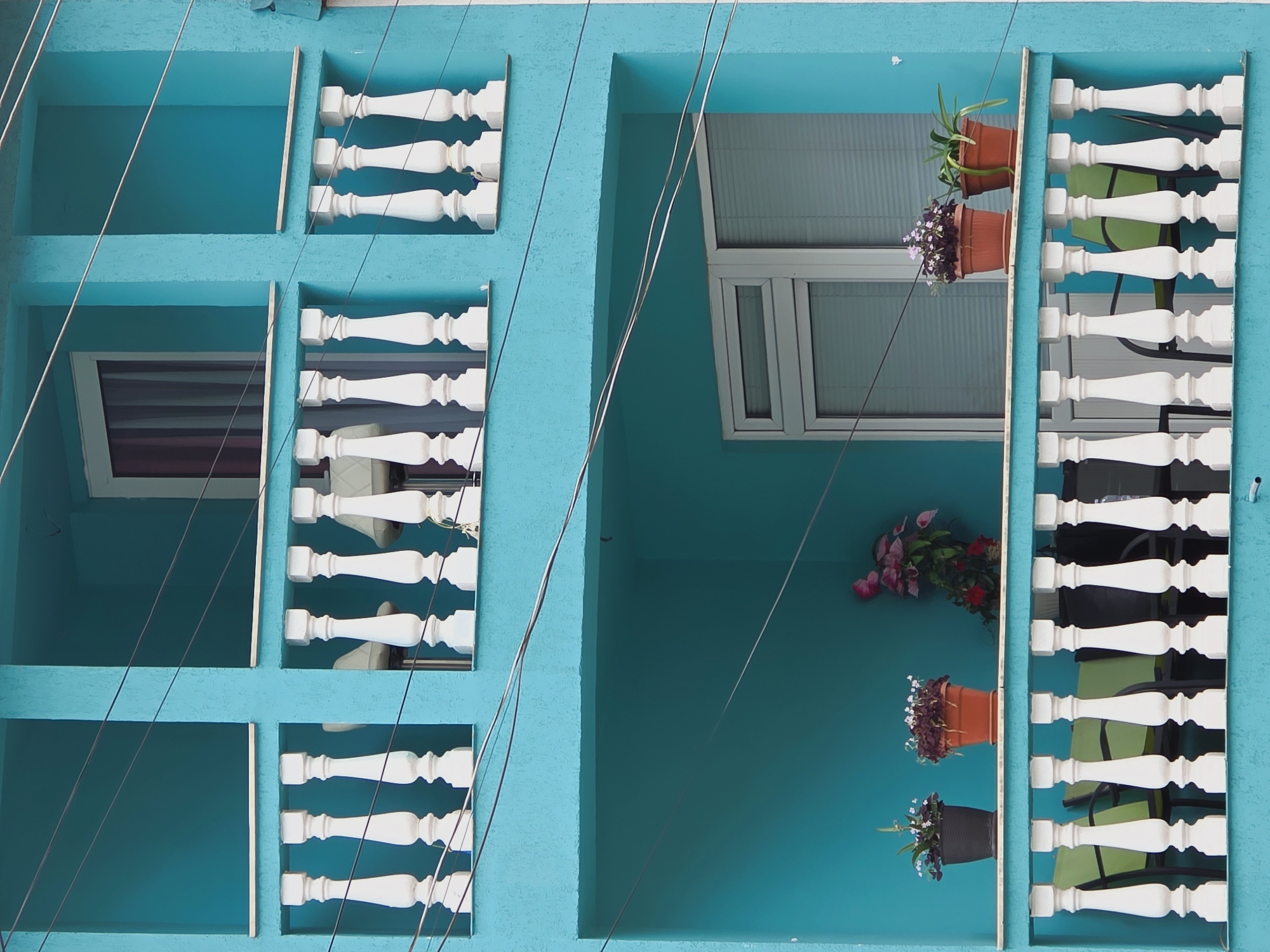 The image shows a close-up of a bright turquoise building with balconies in Ribnovo, Bulgaria. The building has two levels of balconies, each with white balustrades. The upper balcony has two window openings, one with a window with curtains and another empty space. The lower balcony features a window and several potted plants. On the lower balcony, to the left, there are two potted plants with small white flowers in dark pots, and to the right, a larger arrangement of pink and red flowers. In front of the lower balcony railing, there are several green chairs. Power lines are visible crisscrossing the front of the building.