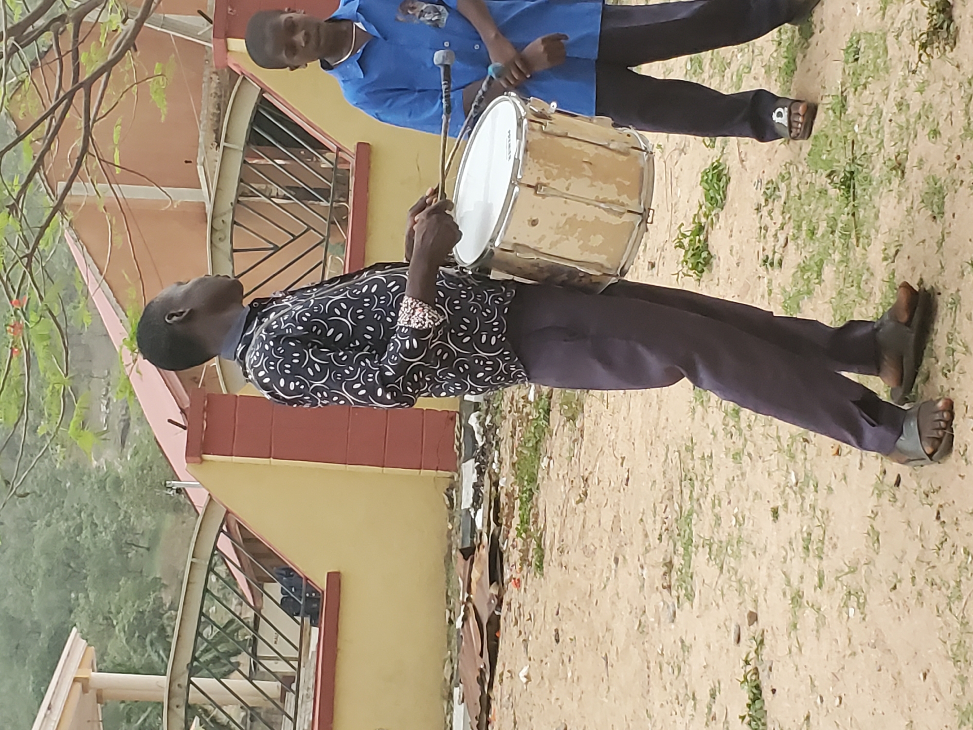 A man wearing a patterned shirt and dark pants stands on a dirt and grass surface, holding and preparing to play a white and tan bass drum with drumsticks. Another man in a blue shirt and dark pants stands behind him, holding a microphone. The scene appears to be outdoors, next to a tan and red building with barred windows. The location is Mararaba, Nigeria.