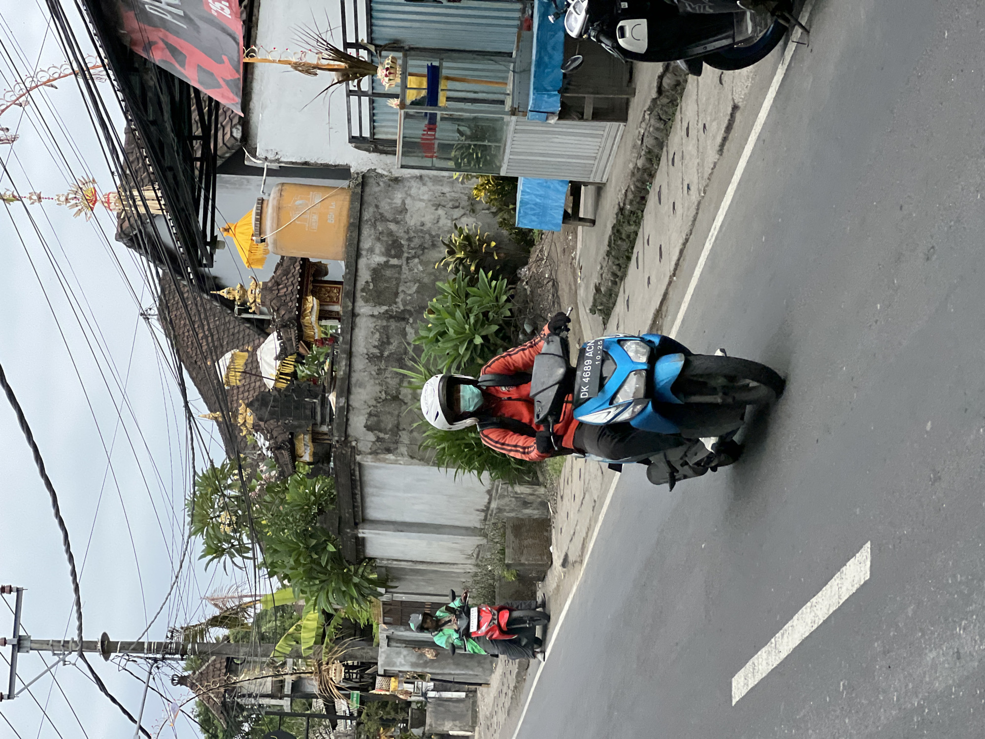 The image depicts a street scene in Denpasar, Indonesia, under an overcast sky. Two individuals are riding scooters on a paved road. The scooter in the foreground is blue, and the rider wears an orange jacket, black pants, gloves, a white helmet, and a light blue face mask. The scooter behind them is red, and the rider is wearing a green jacket and a dark helmet. Both scooters are moving forward.
The roadside features a concrete wall with embedded planters containing green foliage. Behind the wall, traditional Balinese architecture is visible, with intricately decorated rooftops and structures adorned with gold accents. Electrical wires are strung overhead, supported by a utility pole. Several small shops or stalls with blue tarpaulins are set up along the sidewalk. In the background, a large yellow water tank is mounted on a structure. A black scooter is parked on the right side of the road, partially visible. The overall impression is that of a typical urban street in Bali, characterized by its unique cultural elements and dense utility infrastructure.