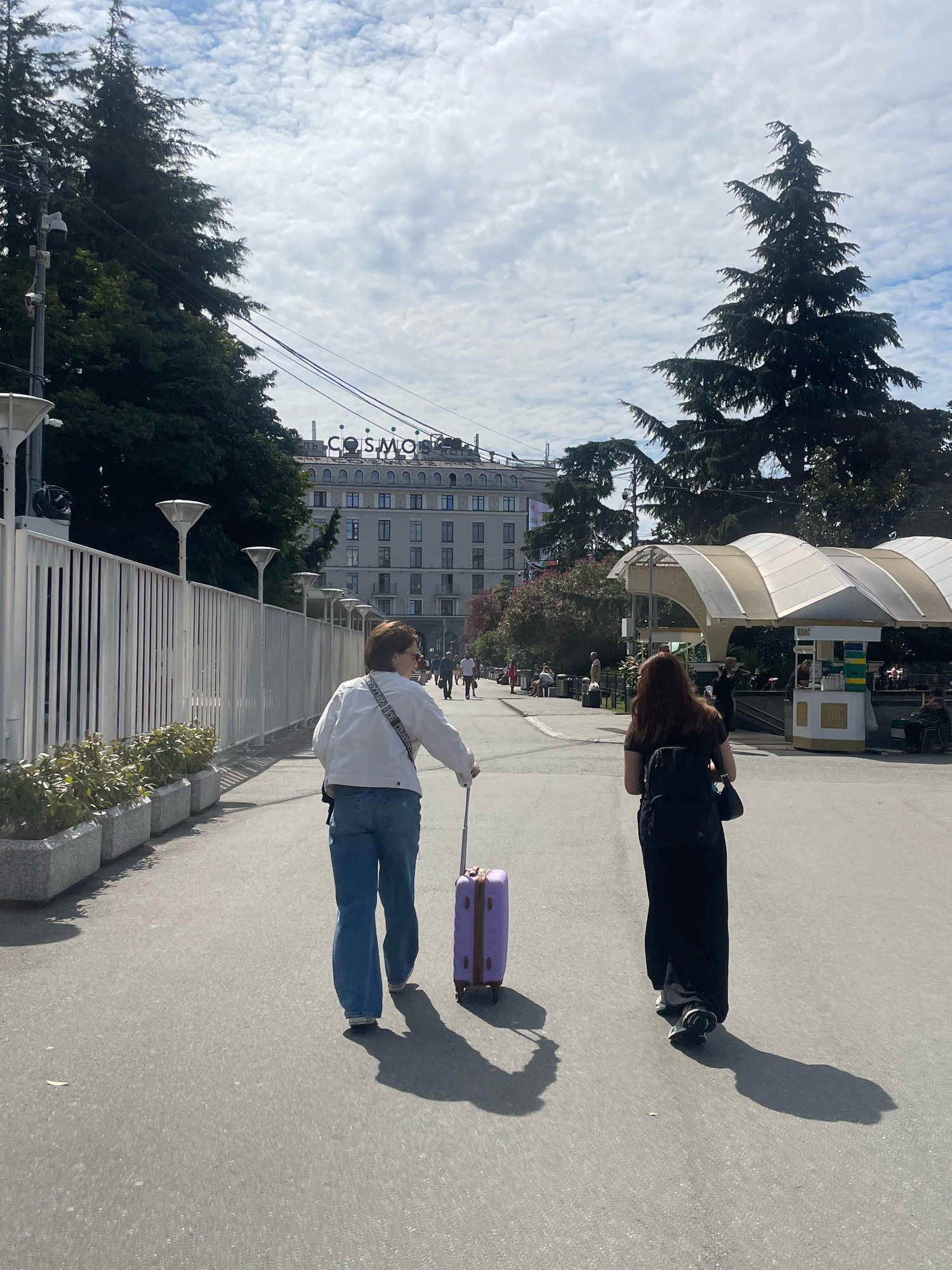 Two women walk on a paved pathway. The woman on the left wears a white jacket and blue jeans and pulls a purple suitcase. The woman on the right wears a black dress and a black backpack. They are walking towards a building with "COSMOS" written on it. The scene appears to be in Sochi, Russia, possibly near a hotel or resort, given the architecture and vegetation. The sky is cloudy and blue.