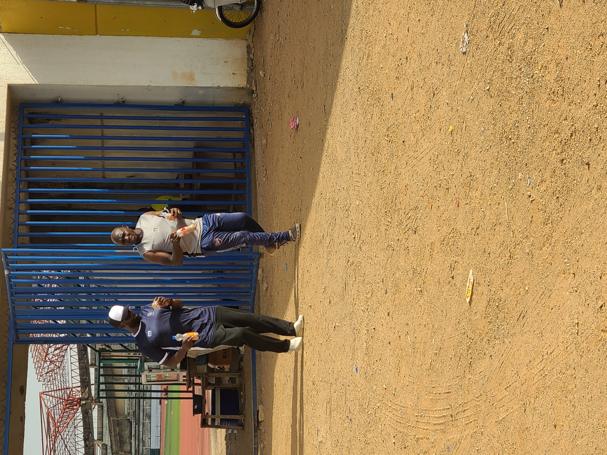 The image shows two men standing near a blue metal gate next to a dirt ground at New Stadium, Nigeria. The man closer to the viewer wears a dark blue shirt, black pants, and white shoes, holding a bottle and what appears to be a small snack. The other man is wearing a light grey t-shirt, dark blue sweatpants, and also holds a bottle. In the background, the stadium’s red metal structure can be seen alongside the green turf field. A wall is behind the men and the gate and the image is rotated.