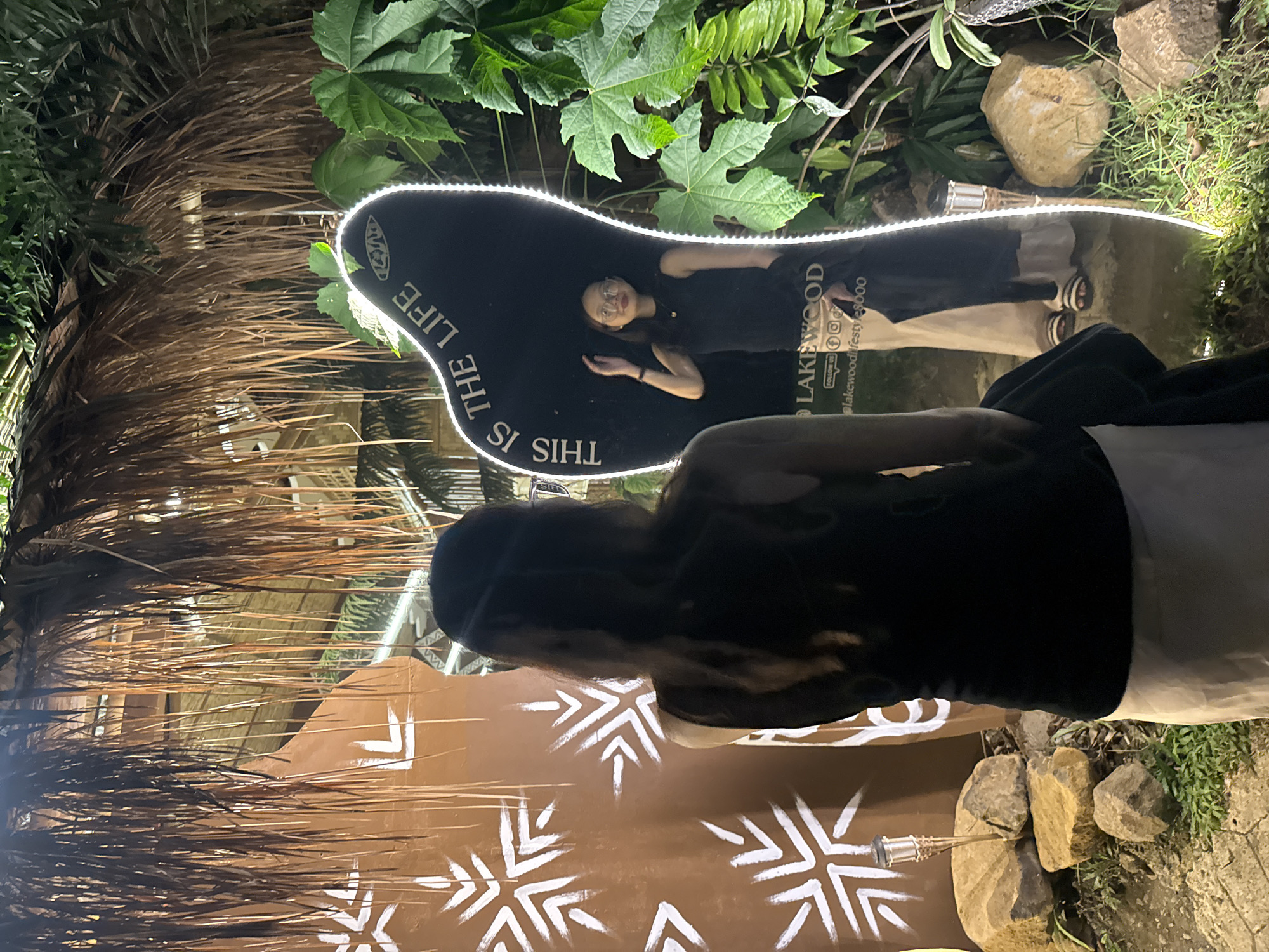A dark-haired woman wearing a black shirt and light-colored pants stands in front of a mirror shaped like a footprint with the words "THIS IS THE LIFE LAKEWOOD" printed in white with a lighted border. The reflection in the mirror shows a woman with glasses wearing a dark dress. The area around the mirror is decorated with plants, rocks, and a brown wall with white snowflake-like designs. The scene appears to be outdoors in Davao City, Philippines.