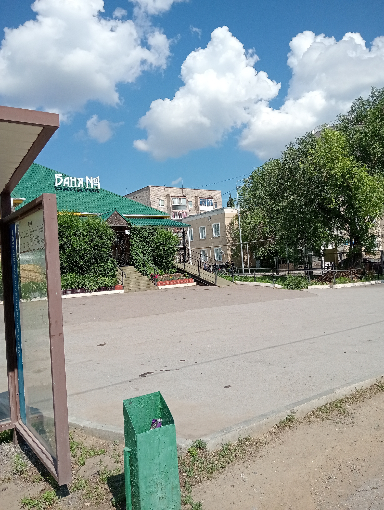 The image depicts an outdoor scene on a sunny day. In the foreground, a green trash can is visible, containing some purple litter. To the left, part of a bus stop shelter with a glass panel is present. In the midground, a building with a green, sloped roof is prominently featured, with the sign "Баня №1" (Banya No. 1) displayed on its facade. The building is surrounded by lush green bushes and trees. A ramp leads up to the building's entrance, which has a dark-colored door. In the background, several other buildings are visible, along with mature trees. The ground is paved with asphalt and concrete. The sky is blue with fluffy white clouds.