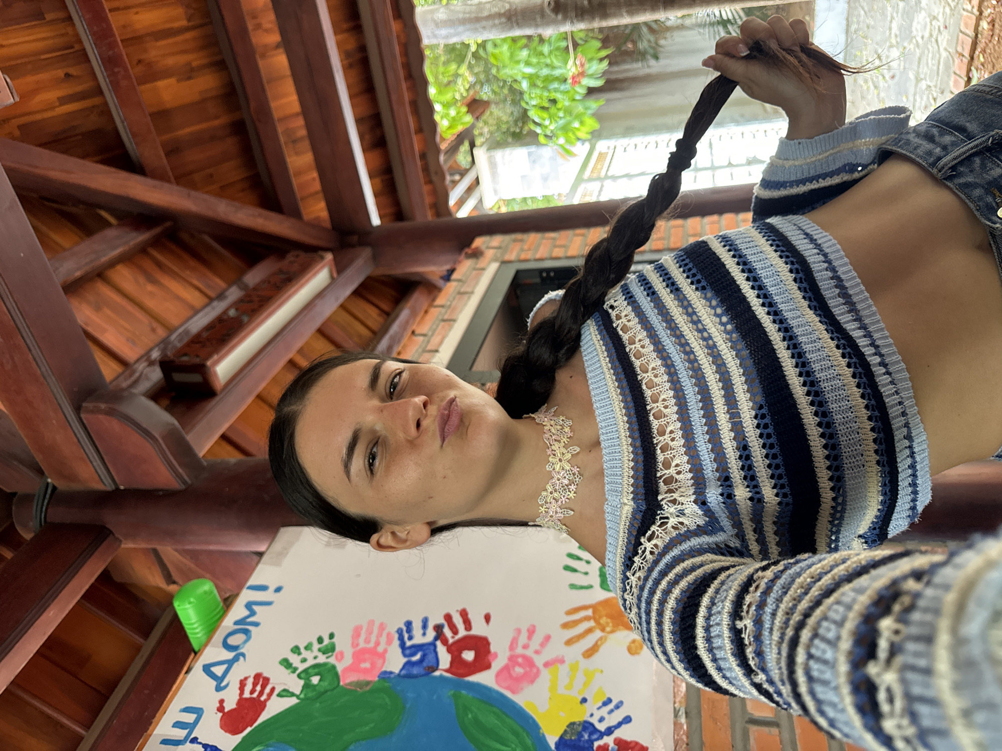 A young woman with dark hair braided down her back is posing for a selfie outdoors, possibly in a covered patio area. She is wearing a cropped, striped knit sweater in shades of blue, white, and beige, and denim jeans. A delicate, floral choker necklace adorns her neck. Behind her, a white board displays colorful handprints and text that appears to be in Russian, with the words "Ш ДОМ!" visible. The background shows a wooden roof structure and lush green foliage, consistent with a tropical setting. The scene suggests a casual, possibly educational or community-oriented environment.