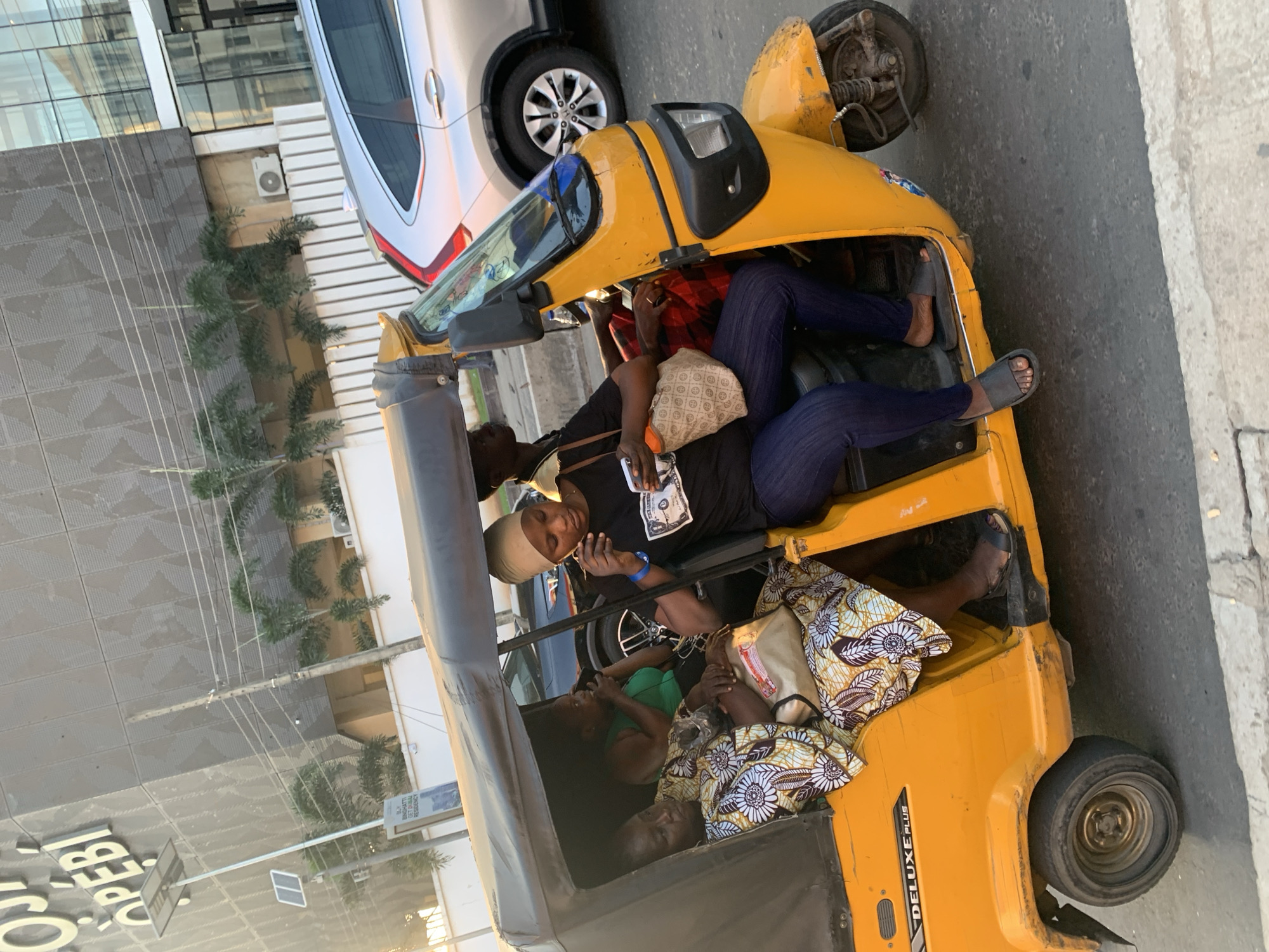 A bright yellow three-wheeled auto-rickshaw, identified by the "DELUXE PLUS" lettering on its side, is captured in motion on a street in Ikeja, Nigeria. The auto-rickshaw carries four individuals. In the driver's seat, a person with dark skin wears a black t-shirt and dark blue pants, holding a mobile phone and looking towards it. Beside the driver, another person with dark skin, wearing a green tank top, is partially visible and appears to be speaking. In the rear passenger section, a woman with dark skin is seated, dressed in a vibrant yellow and white patterned dress and carrying a beige handbag. Another woman with dark skin is positioned in front of her, also in a patterned dress. The background reveals a modern building with a grey, textured facade, palm trees, and a silver SUV. The lighting suggests it is daytime, possibly late afternoon, with soft sunlight illuminating the scene. The street appears to be paved, with a white curb visible in the foreground.