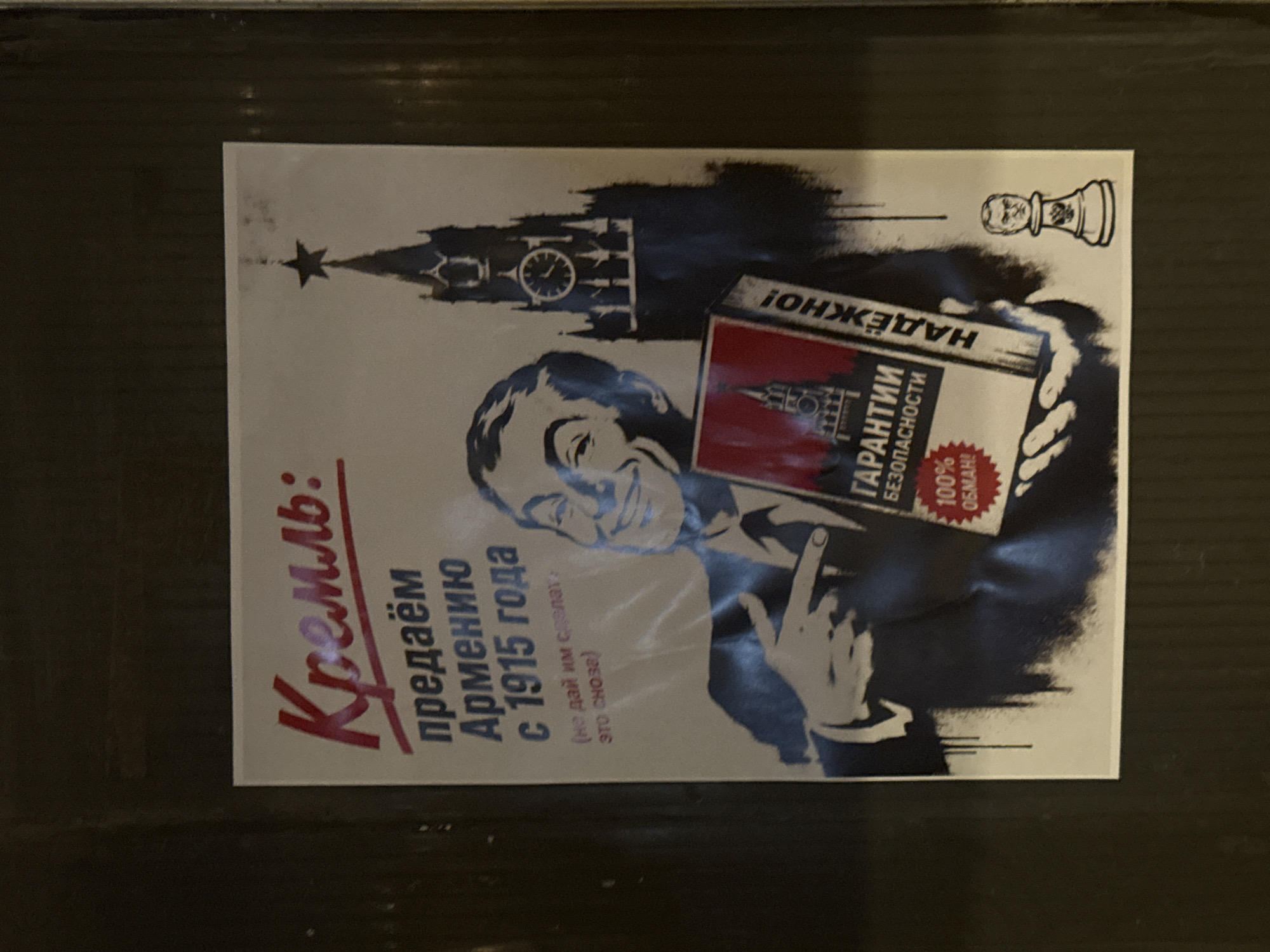 The image displays a political poster or stencil art, likely affixed to a dark, textured surface, in Yerevan, Armenia. The artwork features a caricatured depiction of a man, possibly a political leader, with a slight, knowing smile. He is holding a box labeled with text. Above him, a silhouette of the Spasskaya Tower of the Moscow Kremlin is visible, topped with a star and featuring its iconic clock face. A chess piece, resembling a king or pawn, floats in the upper right corner of the poster.
The poster employs a stark black and white color scheme, with prominent red accents used for emphasis on certain text and the box. The man's gesture of presenting the box, combined with the overlaying text, suggests an activity of offering or propaganda, framed critically by the poster's message.
Visible text on the poster is primarily in Russian. On the left side, large red text declares "Кремль: предаём Армению с 1915 года" (Kremlin: betraying Armenia since 1915). Beneath this, smaller text in parentheses reads "(не дай им сделат это снова)" (don't let them do it again). The box held by the man features the words "ГАРАНТИИ БЕЗОПАСНОСТИ" (GUARANTEES OF SECURITY) in white on a red background, with "100% ОБМАН!" (100% DECEIT!) written below it. Additionally, "НАДЁЖНО!" (RELIABLE!) is written upside down on what appears to be the lid of the box. The poster critiques Russian policy toward Armenia, referencing historical events and implying false promises of security.