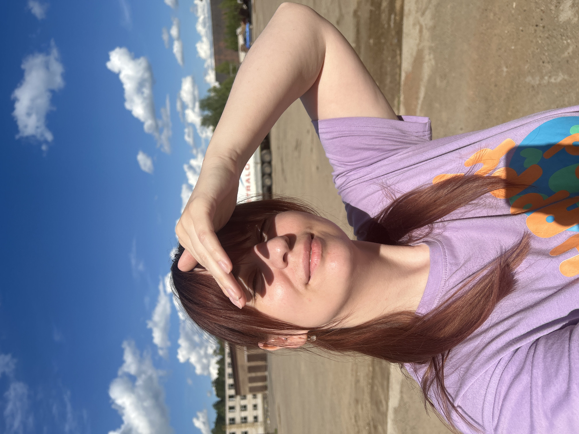 A person with reddish-brown hair and a lilac t-shirt is holding their hand above their brow, possibly to shield their eyes from the sun. The sky is bright blue with white clouds. In the background, there appears to be a building, possibly an apartment block. There is a truck and other vehicles behind the person. The ground is a light shade of brown. The picture was taken in the city of Chaikovsky, Russia.