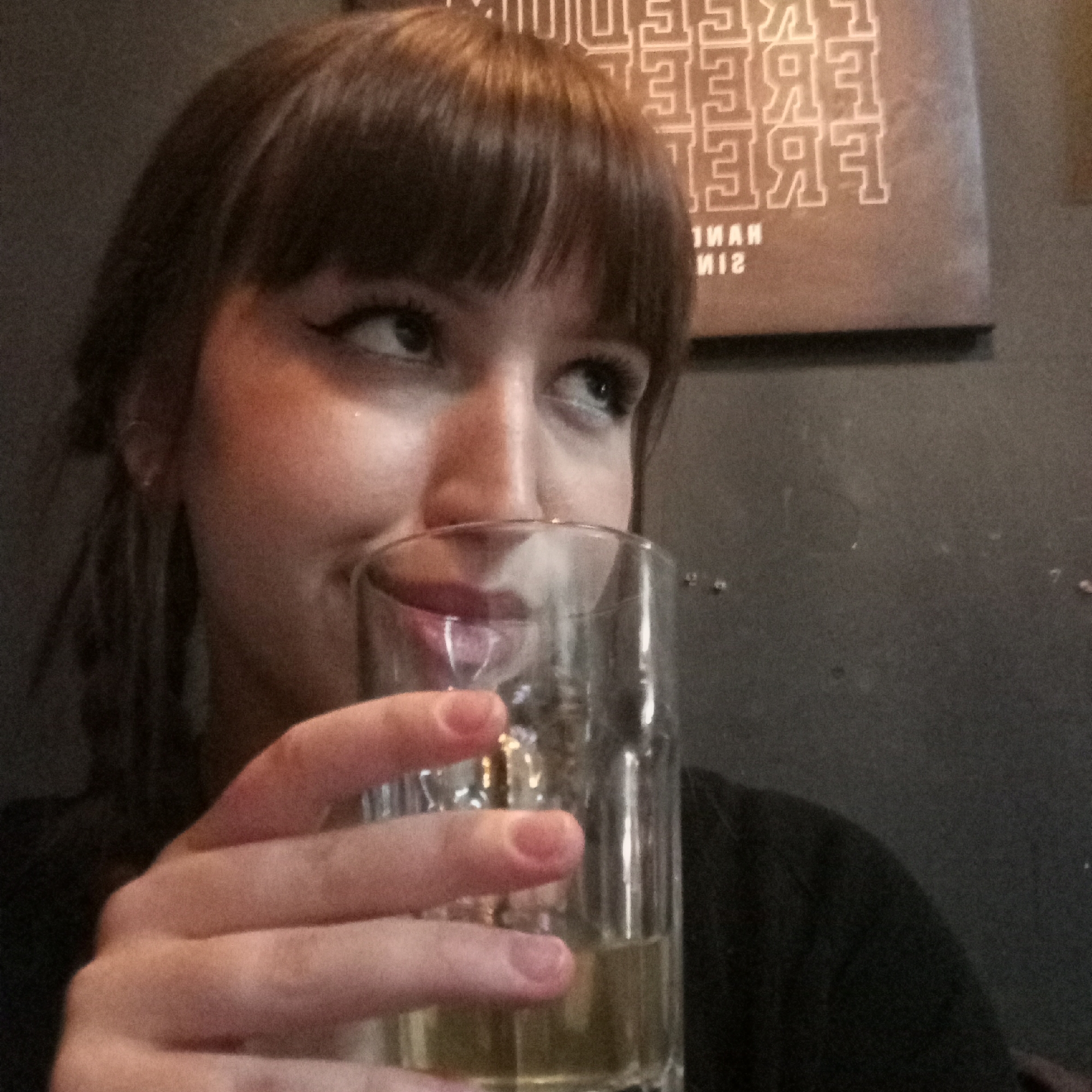 A fair-skinned woman with dark hair and bangs is holding a glass containing a yellow-tinted beverage. The woman is looking off to the side, and the rim of the glass is near her mouth. Her fingers are prominently displayed on the glass.
Behind the woman, a wall is visible. A framed print or poster with stacked text that reads "FREE" in a stylized manner can be seen partially in the background.
The exact location within the city of Chaikovsky, Russia, cannot be determined from the visual information provided.
