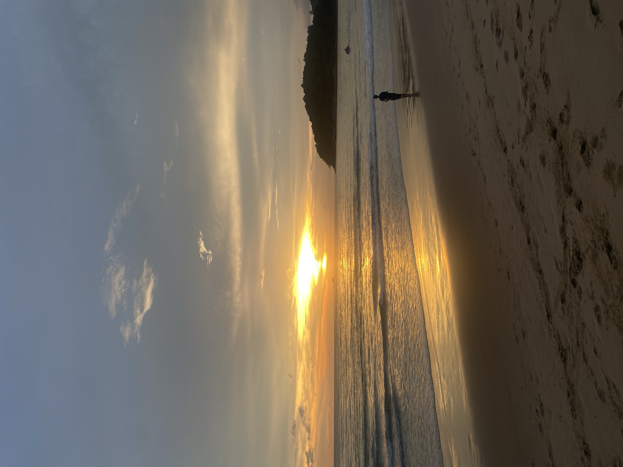 The image shows a sunset scene on a beach, likely in Choeng Thale, Thailand, given the prompt. A person stands in the shallow water at the edge of the beach, silhouetted against the bright sunlight reflecting on the wet sand and waves. To the left, the sky is a gradient of blue to orange and yellow where the sun is setting. Light cloud formations are scattered across the sky. A tree-covered landmass is visible in the background, extending along the coastline. The sand is pale and marked with footprints. The image is rotated counter-clockwise.