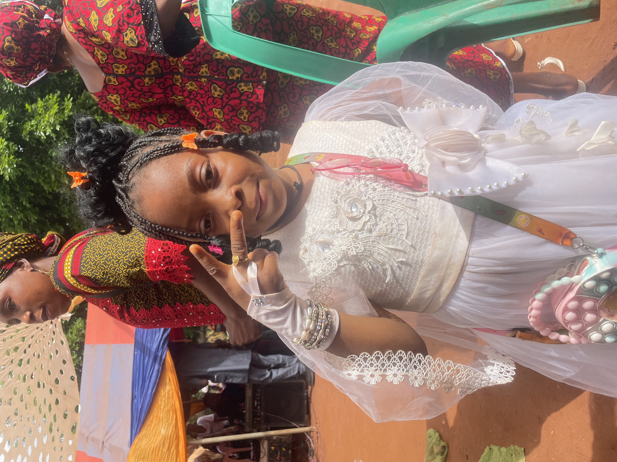 In Ogbe, Nigeria, a young girl in a white dress poses, holding up a peace sign. Her hair is styled in braids with orange butterfly clips. She wears white gloves with lace trim and several bracelets on her left wrist. A pink and blue beaded purse hangs off her right shoulder. Behind her, two women are partially visible. One wears a red, yellow, and black patterned dress and head wrap. The other woman also wears a patterned top. A green plastic chair is also visible in the background. In the lower portion of the frame, a blue, white, and orange tent-like structure is seen, with glimpses of equipment behind it. The ground is reddish-brown.