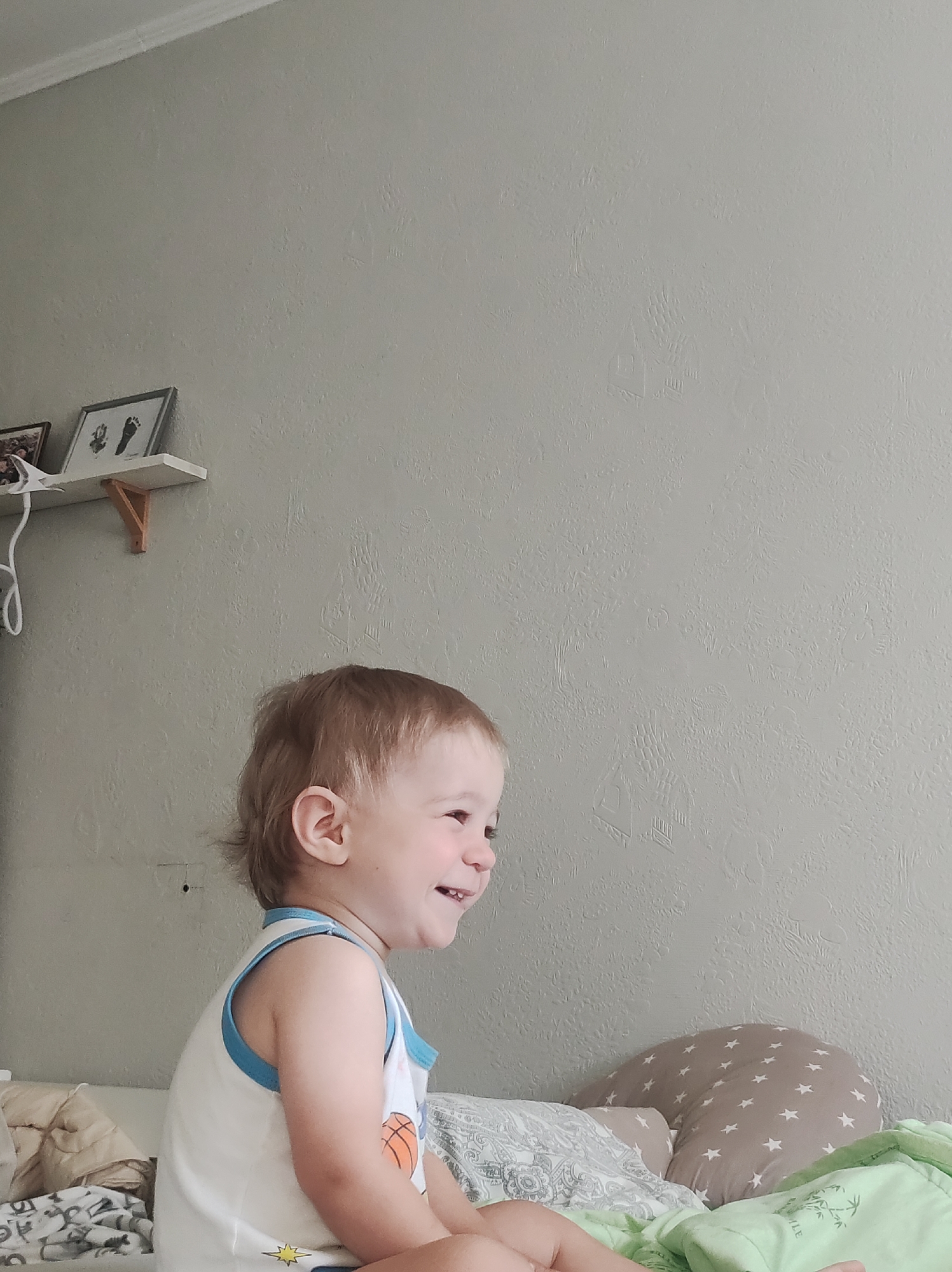 The image captures a cheerful toddler, likely a boy, sitting indoors, possibly in a bedroom or nursery in Chaikovsky, Russia. The child, with light brown hair and a fair complexion, is seen from the chest up in profile, facing right and smiling broadly, with laughter evident in his expression. He is wearing a white sleeveless top with blue trim, featuring an orange basketball graphic and a yellow star design at the bottom.
The background consists of a light gray wall with a textured finish. In the upper left quadrant, a white floating shelf supported by a small wooden bracket is visible. On the shelf, there are two framed pictures; one clearly displays black footprints, typical of a baby's keepsake. A white coiled cord hangs down from the shelf. To the right of the child, a patterned pillow with white stars on a beige background is partially visible, along with some green fabric, which has the partial text "TLE" printed on it, possibly part of the word "LITTLE." Other bedding or fabric with white and gray patterns can be seen in the lower left and behind the child. The lighting appears bright and even, suggesting daytime. The overall atmosphere conveyed is one of joy and innocence.
