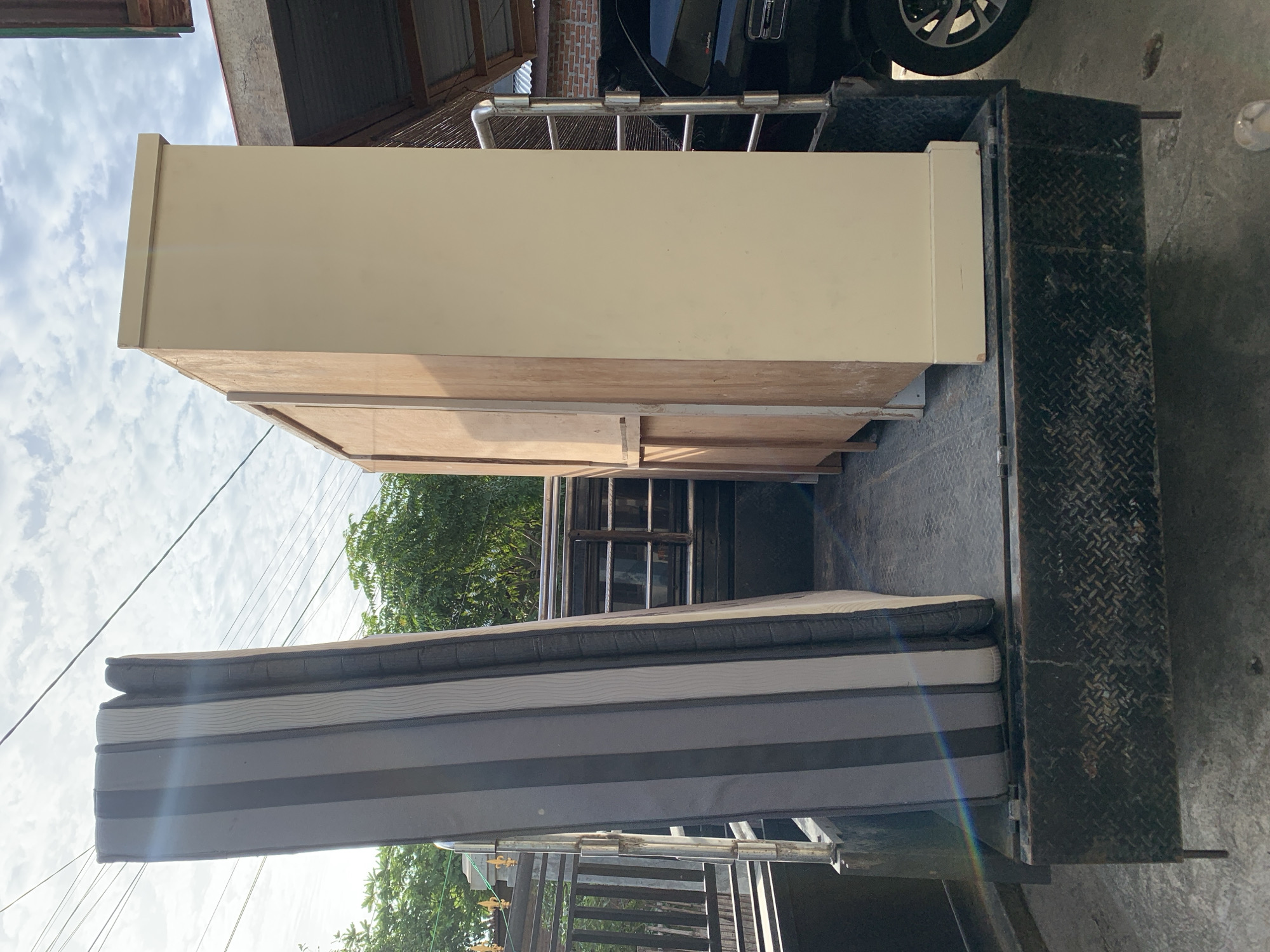 In the city of Meunasahdrang, Indonesia, a truck bed is loaded with furniture. A striped mattress, predominantly gray and white, stands upright on the left side of the truck bed. To its right, a tall, light-colored cabinet with unfinished wood accents is positioned. The truck appears to be parked outdoors on a concrete surface, with the sky visible above. In the background, greenery from trees and a building with a corrugated metal roof are present. A portion of a black vehicle is visible to the right of the cabinet, with its wheel showing. Electrical wires crisscross the sky.