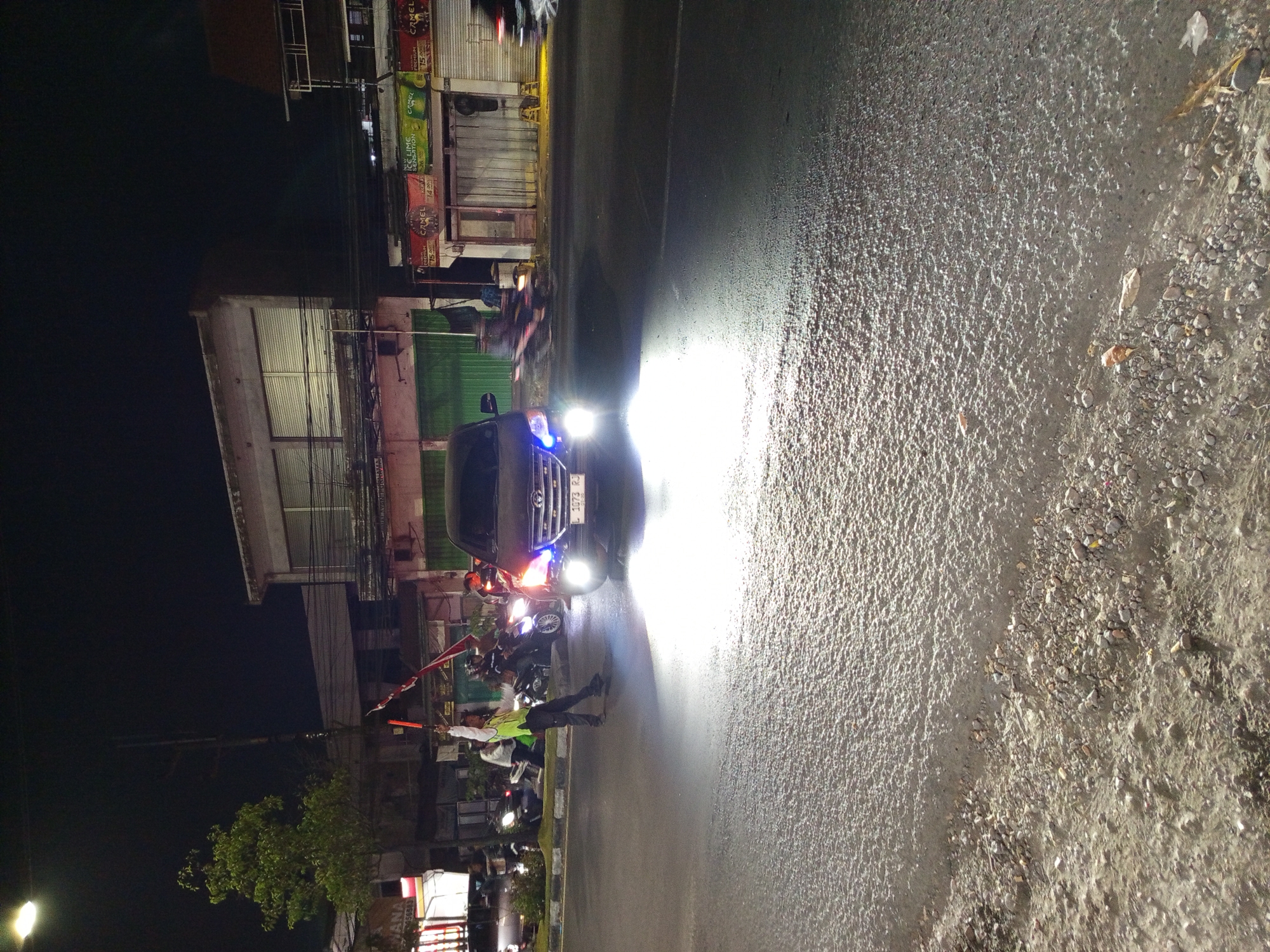 A person wearing a high-visibility vest and holding two illuminated signal batons directs traffic on a wet road at night in Buduran, Indonesia. The person is positioned near the center of the road, illuminated by the headlights of a dark-colored vehicle approaching from the opposite direction. The vehicle's license plate reads "L 1073 RJ". Several motorcycles are visible on the roadside and in the background, some with illuminated headlights. Buildings line the street, with storefronts displaying signage, including what appears to be advertisements for "Camel" cigarettes. The road surface is dark and reflects the light from the vehicle's headlights, indicating it may be wet or recently rained on. The overall scene depicts a nighttime traffic management scenario on a street in Buduran.