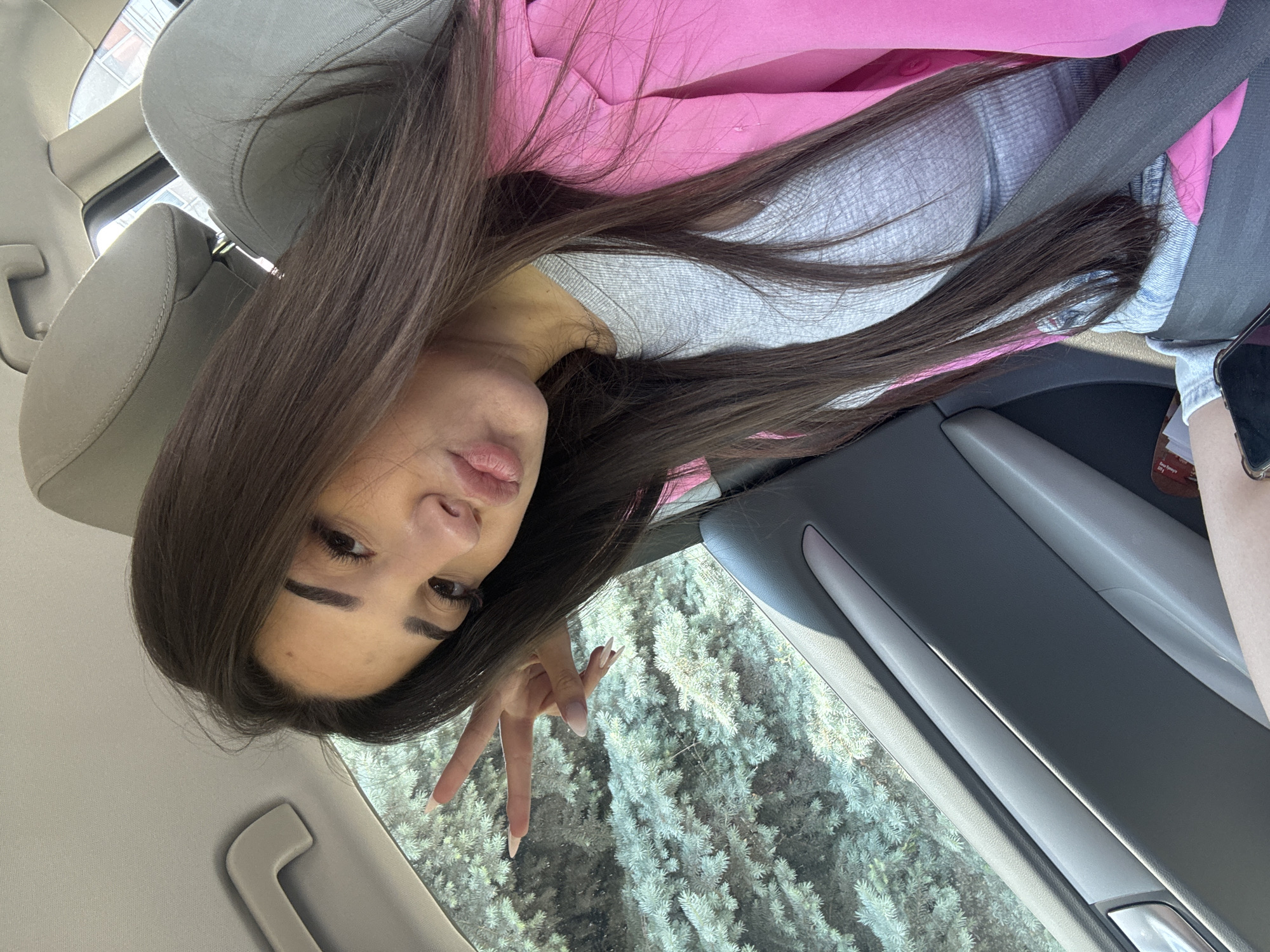 A young woman is captured in a selfie taken inside a car, likely from the passenger seat, with the image rotated 90 degrees clockwise. She has long, dark brown hair that falls over her shoulders and chest. Her eyes are accentuated with dark eyeliner and what appear to be false eyelashes, complemented by well-defined eyebrows. Her lips are slightly parted in a soft pout or small smile. She is wearing a light grey, possibly waffle-knit, long-sleeved top layered under a bright pink jacket or hoodie. A black seatbelt is visible across her chest, indicating she is buckled in. With her left hand raised, she makes a peace sign (V-sign); her long fingernails are manicured in a natural or French style.
The car's interior features light grey fabric seats, including a visible headrest, and grey and light grey plastic door panels with a silver door handle. Outside the window, lush green foliage is visible, appearing to be coniferous trees like pine or spruce, suggesting a natural or suburban setting. The bright, natural light indicates it is daytime, possibly sunny or partly cloudy. Partially visible near her hand and the car door is a mobile phone and a brown snack wrapper, on which the text "Bear Paw" and "200 g" can be discerned. The overall mood conveyed by her expression and gesture is casual and playful. The location context for this scene is Plovdiv, Bulgaria.