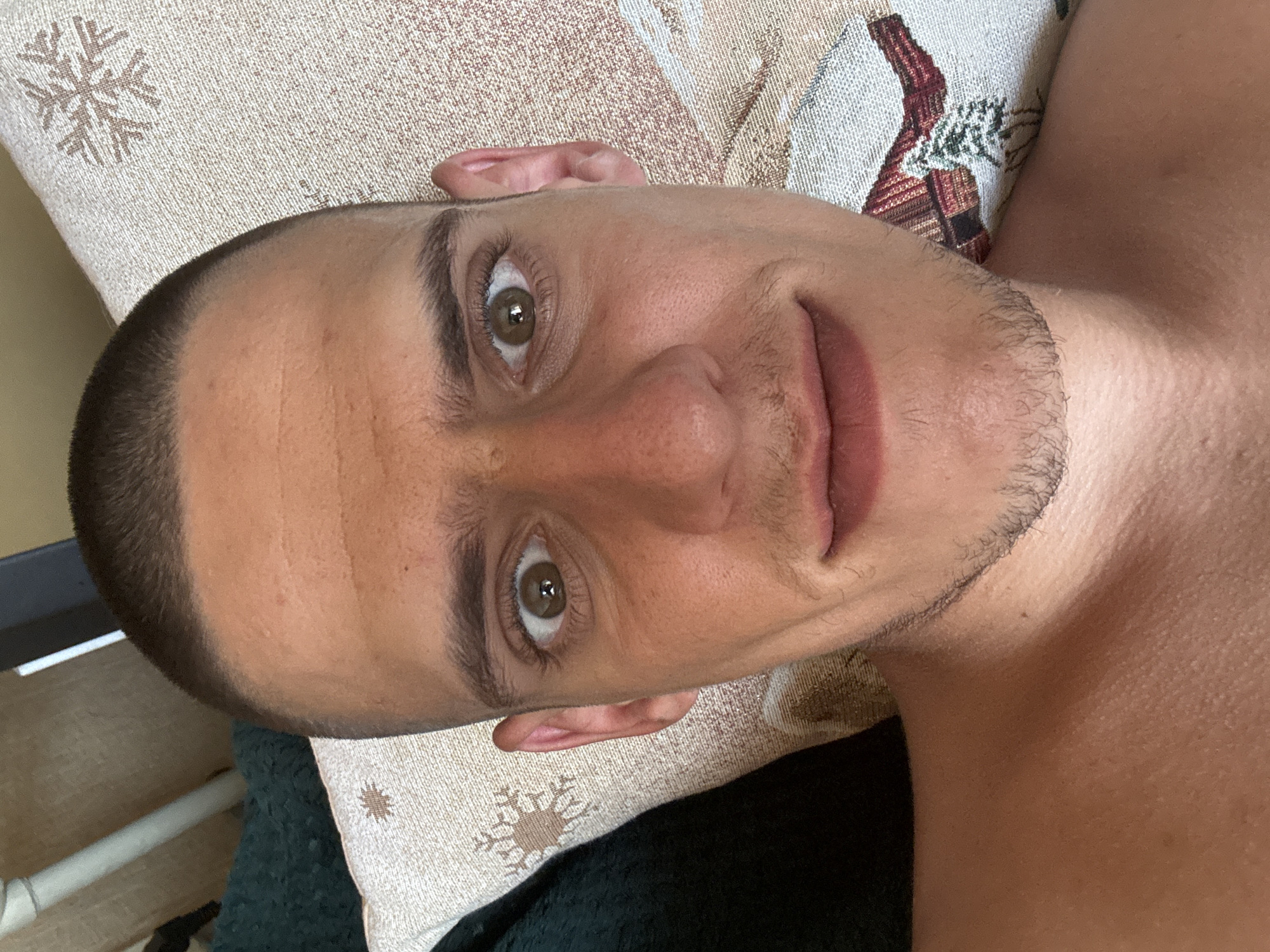 A close-up, overhead selfie shows a man with a buzz cut lying down. He is looking directly at the camera with wide eyes and a slight smile. The man has light brown eyes, dark eyebrows, and stubble on his chin and upper lip. He is shirtless and appears to be indoors, possibly in a living room. Behind him, a textured pillow with a snowflake pattern is visible. The overall lighting suggests daylight. The image does not provide any information or context related to the city of Sofia, Bulgaria.