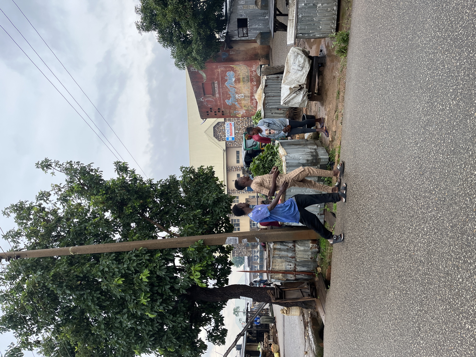The image shows a street scene in Jos, Nigeria, with several people visible. The weather appears overcast, suggesting a cloudy or rainy day.
In the foreground, two young men are standing by a large tree on the side of a paved road. One man, wearing a blue t-shirt and black pants, is in profile, looking towards the other man. The second man, dressed in a patterned light brown outfit, is also in profile and looking in the same direction. Both are wearing sandals. Behind them, a large tree with dense green foliage dominates the left side of the frame, with a utility pole running through its branches.
Further down the road, a group of people are gathered near makeshift stalls constructed from corrugated metal and tarps. Some individuals appear to be selling goods, as suggested by the presence of what looks like produce. A woman in a gray jacket and striped shirt is among them. In the background, there are buildings, some with faded painted signs. One sign is visible on a red-brown building that reads "CAP Private School" and "Tasty Nutrition Grow For You". Another building has a sign with "COCIN" visible. The overall impression is of a bustling, yet somewhat informal, commercial area. The paved road is wet, indicating recent rain.