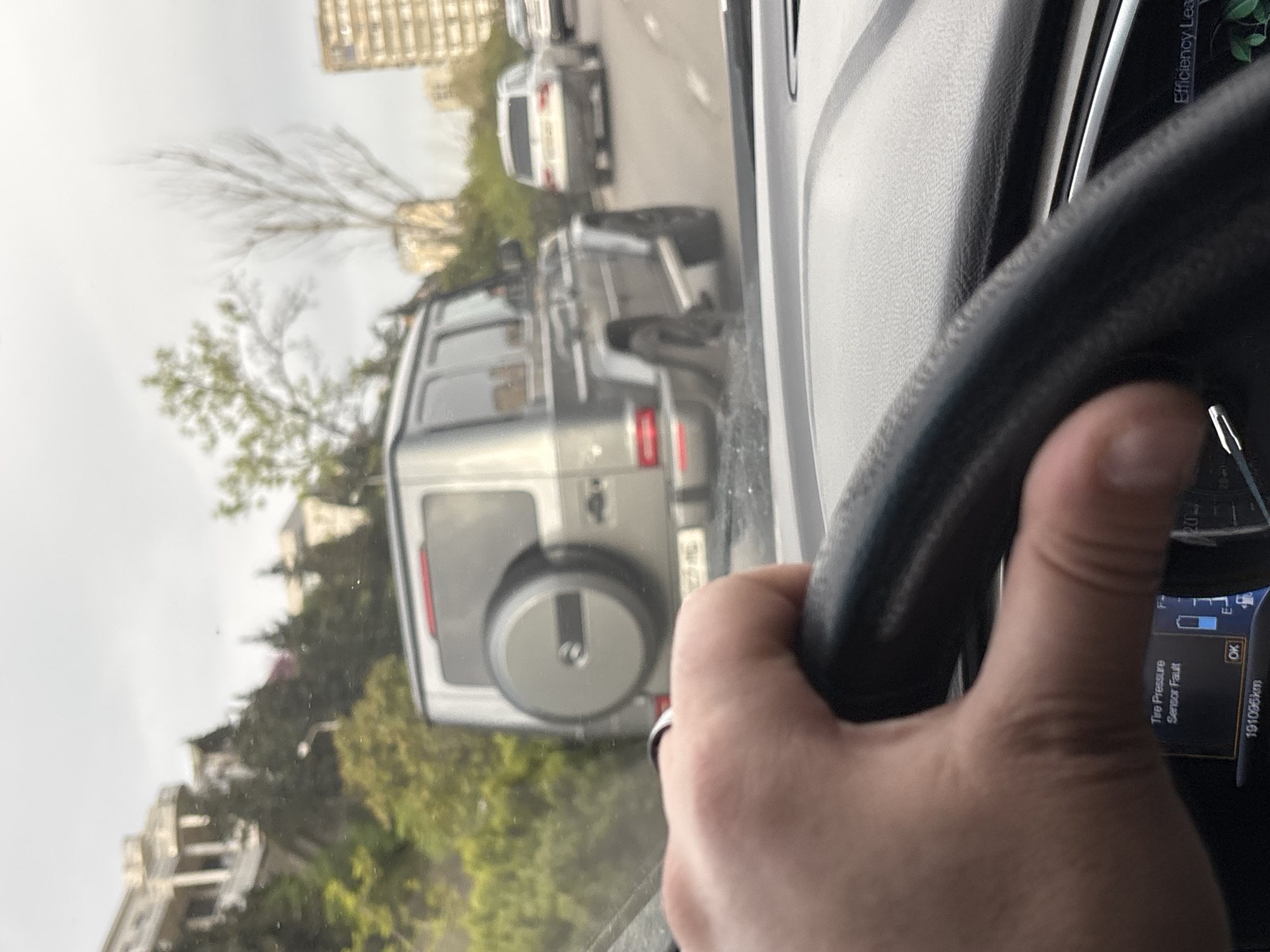 The image presents a first-person perspective from inside a car, looking out through the windshield. A person's right hand is visible, firmly gripping the steering wheel, indicating a driving or passenger situation. The scene unfolds on a road in an urban environment, identified as Tbilisi, Georgia.
Directly in front of the viewer's vehicle is a grey Mercedes-Benz G-Class (G-Wagen), prominently featuring its signature rear-mounted spare tire cover. Further down the road, other cars, including a white SUV, can be seen, suggesting traffic flow or a queue. The background reveals residential or commercial buildings interspersed with trees, some with green leaves and others with bare branches, consistent with an urban setting.
The time of day appears to be daytime, under what looks like an overcast or hazy sky, casting soft, diffused light across the scene.
On the dashboard screen to the right of the steering wheel, several pieces of text are visible: a warning message "Tire Pressure Sensor Fault," an "OK" button, and the vehicle's mileage reading "191096 km." A partial label "Efficiency Lea" is also visible in the upper right of the screen.
