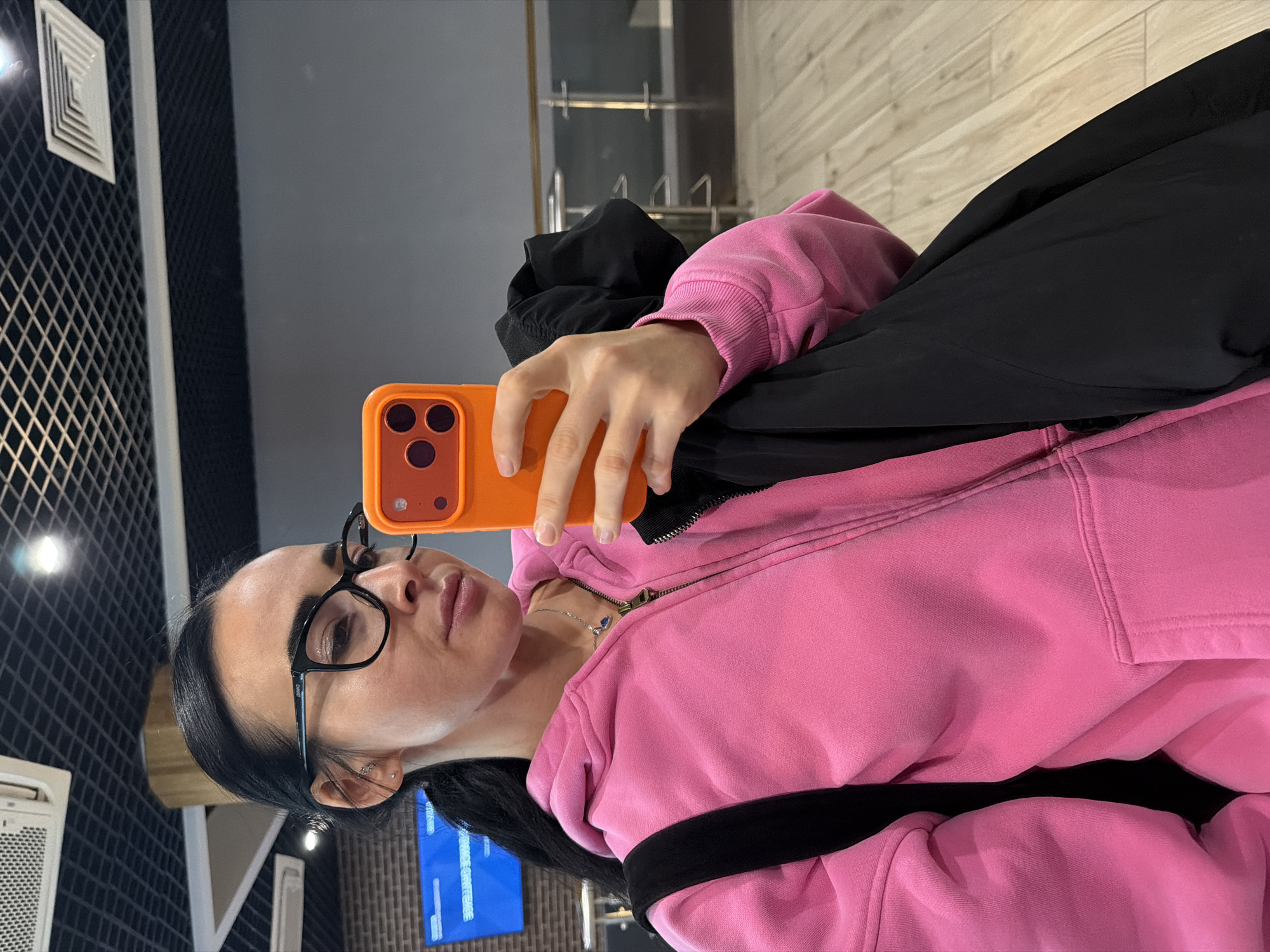 A woman with black hair and glasses is holding an orange smartphone up to take a selfie. She is wearing a pink hooded sweatshirt and a black jacket. The location appears to be an interior space with a black and grey wall design, possibly a public building. A screen with blue text can be seen in the background. This cannot be identified as being specifically in Saint Petersburg, Russia.