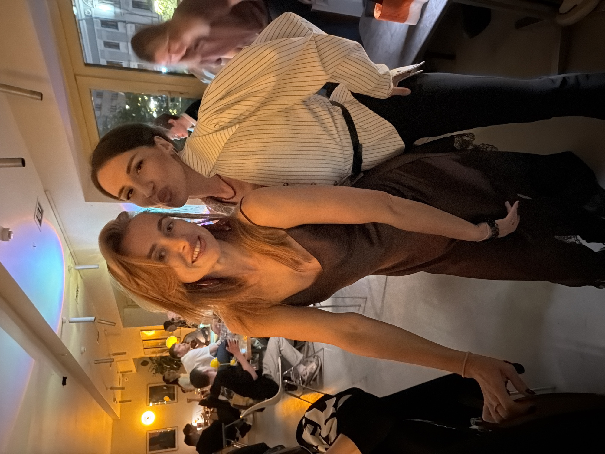 Two women are in the foreground of a horizontally oriented image. One woman wears a brown dress and smiles at the camera. The second woman, behind her, wears a striped button-down shirt. Both women have dark hair. They are located in a crowded restaurant in the city of Krasnoyarsk, Russia. Numerous patrons are seated at tables in the background. The restaurant has warm lighting. A large window is visible in the upper portion of the image, revealing a building and trees outside. The woman in the foreground has her hand on the edge of a table.