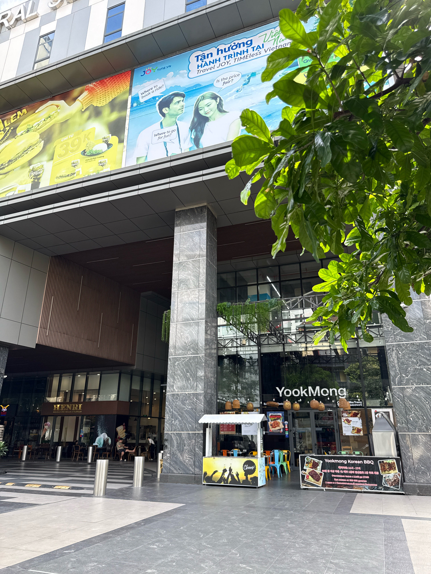 The image depicts the exterior of a modern building in Nha Trang, Vietnam. The building features large glass windows and is adorned with several billboards. One billboard on the left advertises food items like sandwiches and Coca-Cola, with text indicating a price of 39k. Another billboard, to the right of the first, shows a couple and text related to travel and time. A large tree with vibrant green leaves is prominently positioned in the foreground, partially obscuring the view of the building's facade.
At the base of the building, there are storefronts. One storefront labeled "HENRI" appears to be a Belgian ice cream and waffle shop, with tables and chairs visible both inside and outside. Adjacent to this, a Korean BBQ restaurant named "YookMong" is visible, with a prominent sign displaying images of BBQ dishes. A small food stall with colorful chairs is set up outside the YookMong restaurant. Several people are present, some seated at the Henri establishment and others walking or standing near the food stall and the restaurant entrance. The ground in front of the building is paved with large, grey and white tiles. The overall scene suggests a bustling commercial area within the city.
