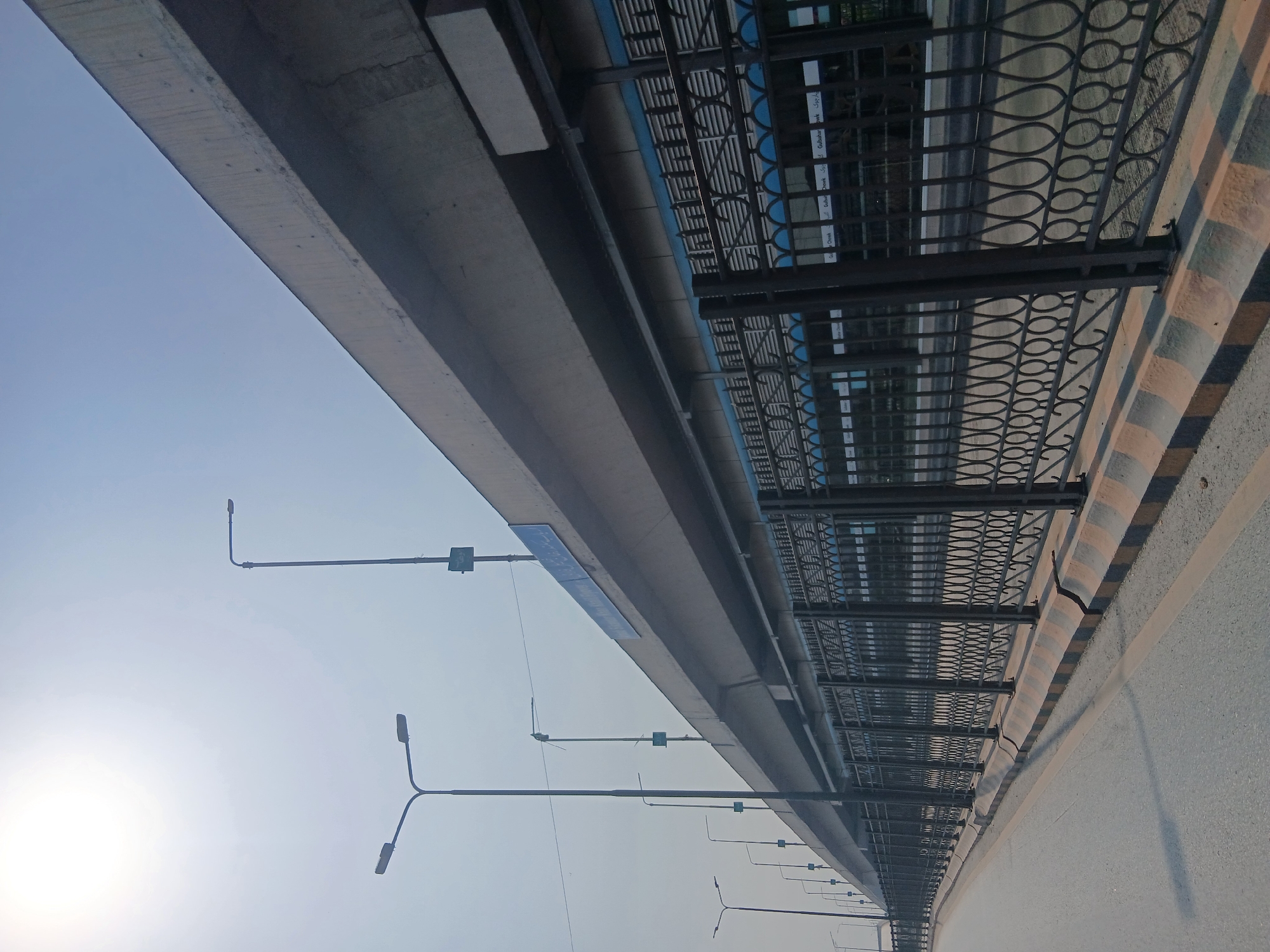 The image shows an elevated road or flyover in Peshawar City Tehsil, Pakistan, on a bright, hazy day. A multi-lane road is visible in the foreground, curving to the left. Along the side of the road is a decorative black metal fence with an ornate pattern. Beyond the fence, the underside of the elevated structure is visible, with a blue and white facade. Several streetlights are positioned along the roadside, and one is mounted on the elevated structure itself. A blue sign with text in Arabic script is attached to the side of the elevated structure. The sky is a pale blue and appears to be washed out by the sun. There are no people visible in the image. The environment suggests an urban or semi-urban setting.