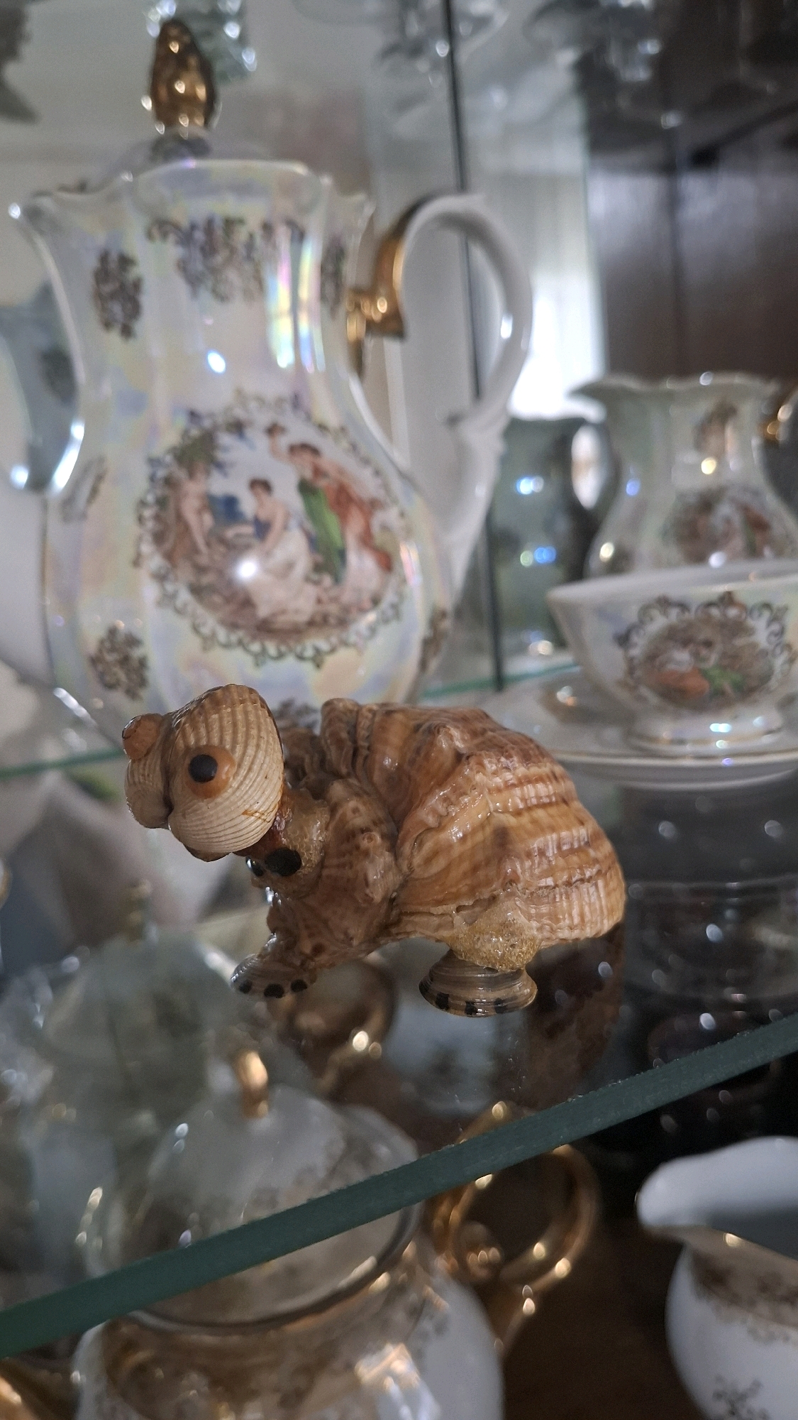 A close-up, eye-level shot showcases a small, decorative turtle figurine made from seashells, positioned in the foreground. The turtle has a shell-like body and head, with small dark beads for eyes and what appear to be tiny beads for its feet. Behind the turtle, a large, ornate pitcher with gold trim and painted scenes of people in historical attire is visible. The pitcher has a pearlescent finish that reflects light, creating a rainbow effect. To the right of the pitcher, a teacup and saucer, also decorated with similar painted scenes and gold accents, are placed on a glass shelf. The items are displayed in what appears to be a glass cabinet, with reflections of lights and the environment visible on the surfaces. The scene is static and appears to be an indoor display of decorative ceramic and shell items. There are no people present in the immediate vicinity of the displayed items. The specific location in Shymkent, Kazakhstan, cannot be determined from the visual information provided in the image.
