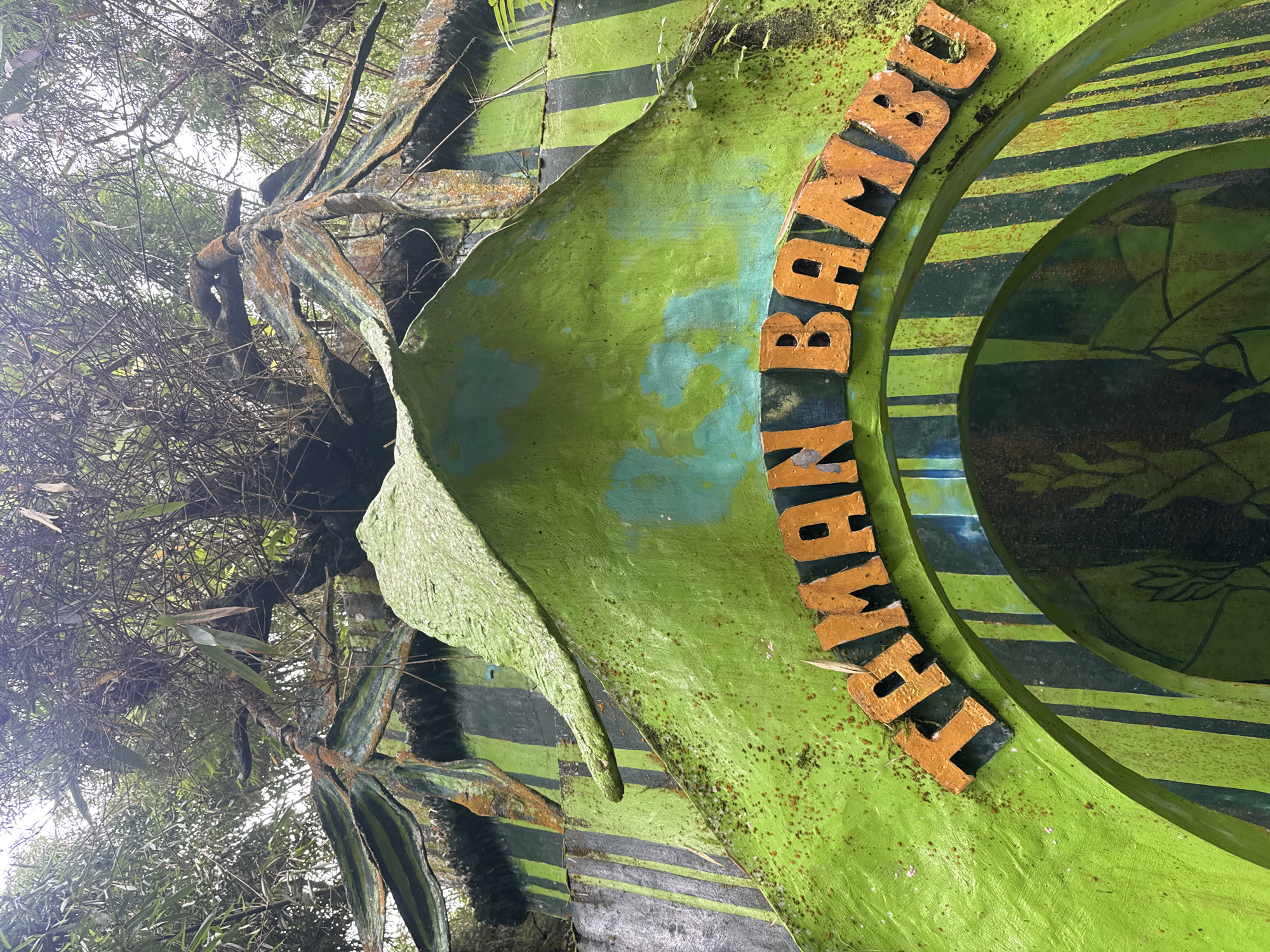 A low-angle shot of the "Taman Bambu" (Bamboo Park) sign in Baturiti, Indonesia, reveals its weathered condition. The sign features green and black stripes, with orange lettering forming an arc. Surrounding the sign is dense vegetation. The lighting suggests overcast conditions, contributing to the subdued atmosphere of the image. The sign appears to be made of plaster and is beginning to crumble in some sections.