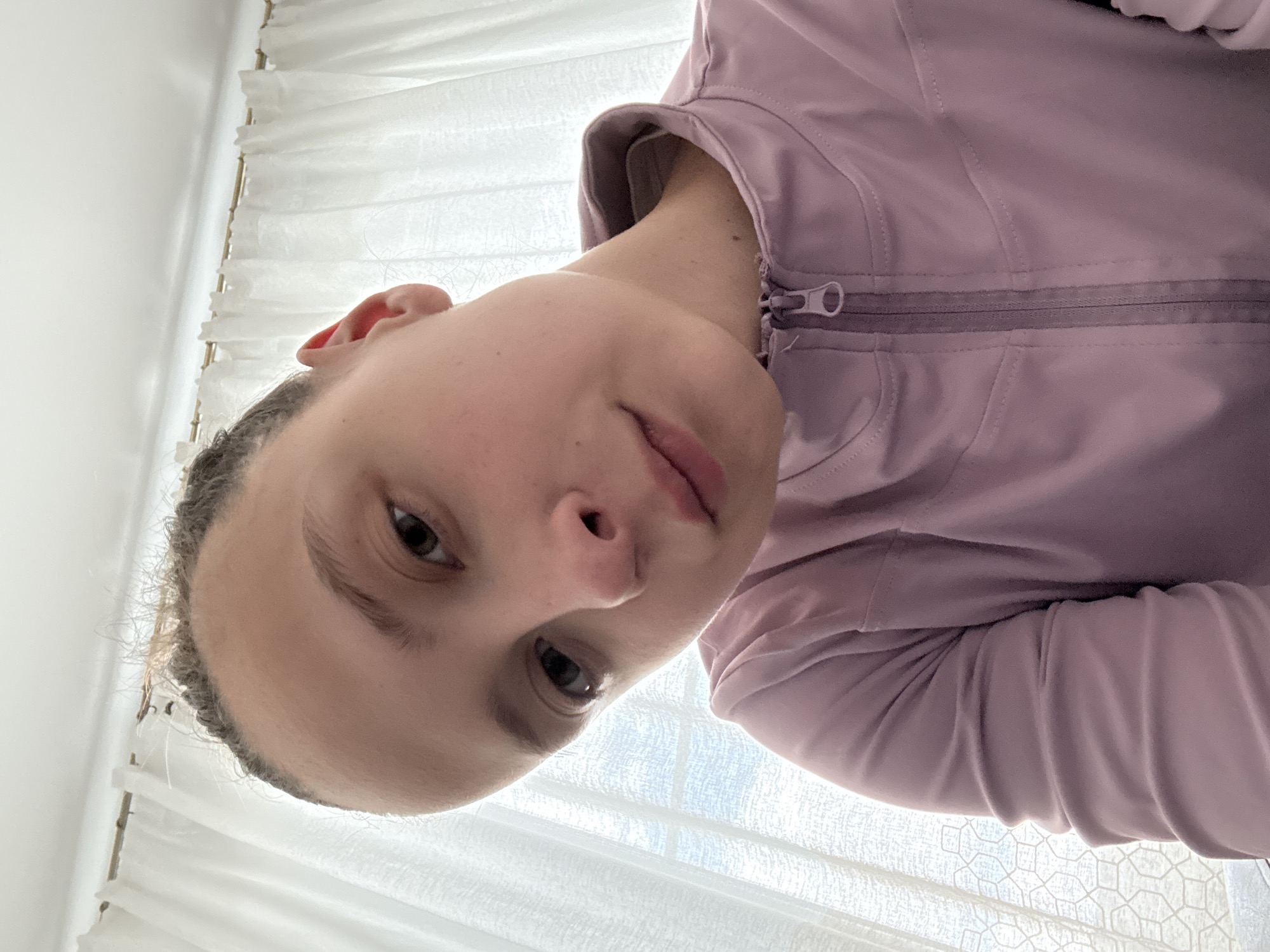 A close-up selfie of a fair-skinned individual with brown hair, cropped short around the head, wearing a zippered, light purple jacket. The person is positioned at an unusual angle, looking down towards the camera. Behind the person is a blurred background of white, sheer curtains. There is no visible information to indicate that the photo was taken in the city of Perm, Russia.