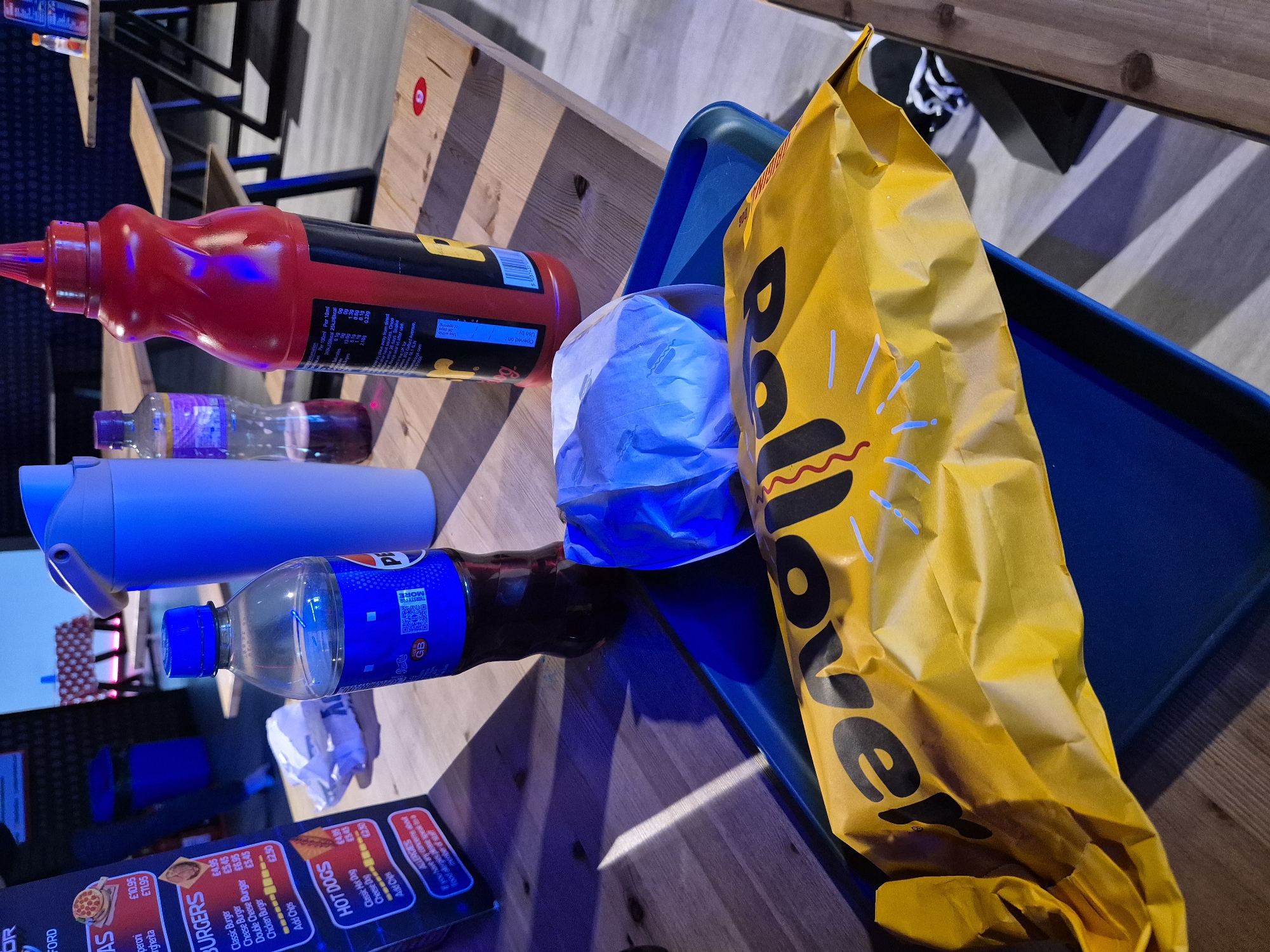 The image captures a tabletop scene featuring a variety of food and drink items, likely in a fast-food or casual dining environment.
Visible items include a large bottle of ketchup, a bottle of an unidentified purple liquid, a blue water bottle, a bottle of Pepsi, a foil-wrapped burger, a bag of food labelled "bollover", and a blue plastic tray. A menu or price list mentioning "Burgers", "Hot Dogs", "Peroni", and "Allergies" is also present.
The backdrop includes hints of chairs and tables, indicating a food establishment. The menu references "Chelmsford" and "Ford Avenue" which implies the location is within the city of Chelmsford, United Kingdom.
