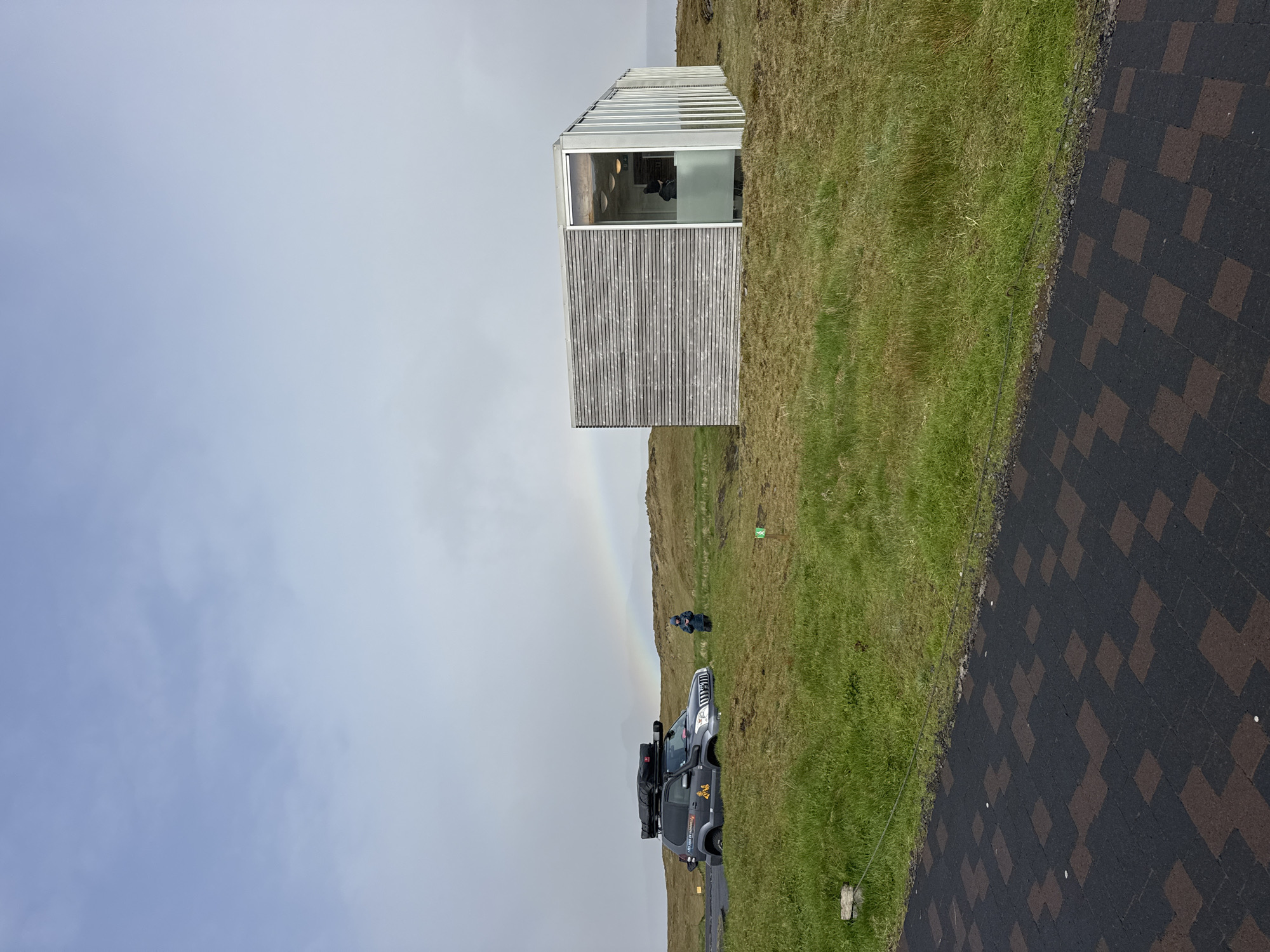 The image shows a landscape in Vik, Iceland, featuring a modern, rectangular building with wooden siding and large windows. The building is situated on a grassy hillside. A partially visible rainbow arcs across the sky. A dark-colored SUV with a roof rack and cargo is parked near the building, and two people are standing near the SUV. A paved road or walkway with dark, patterned tiles borders the grassy area. The sky is overcast and light grey.