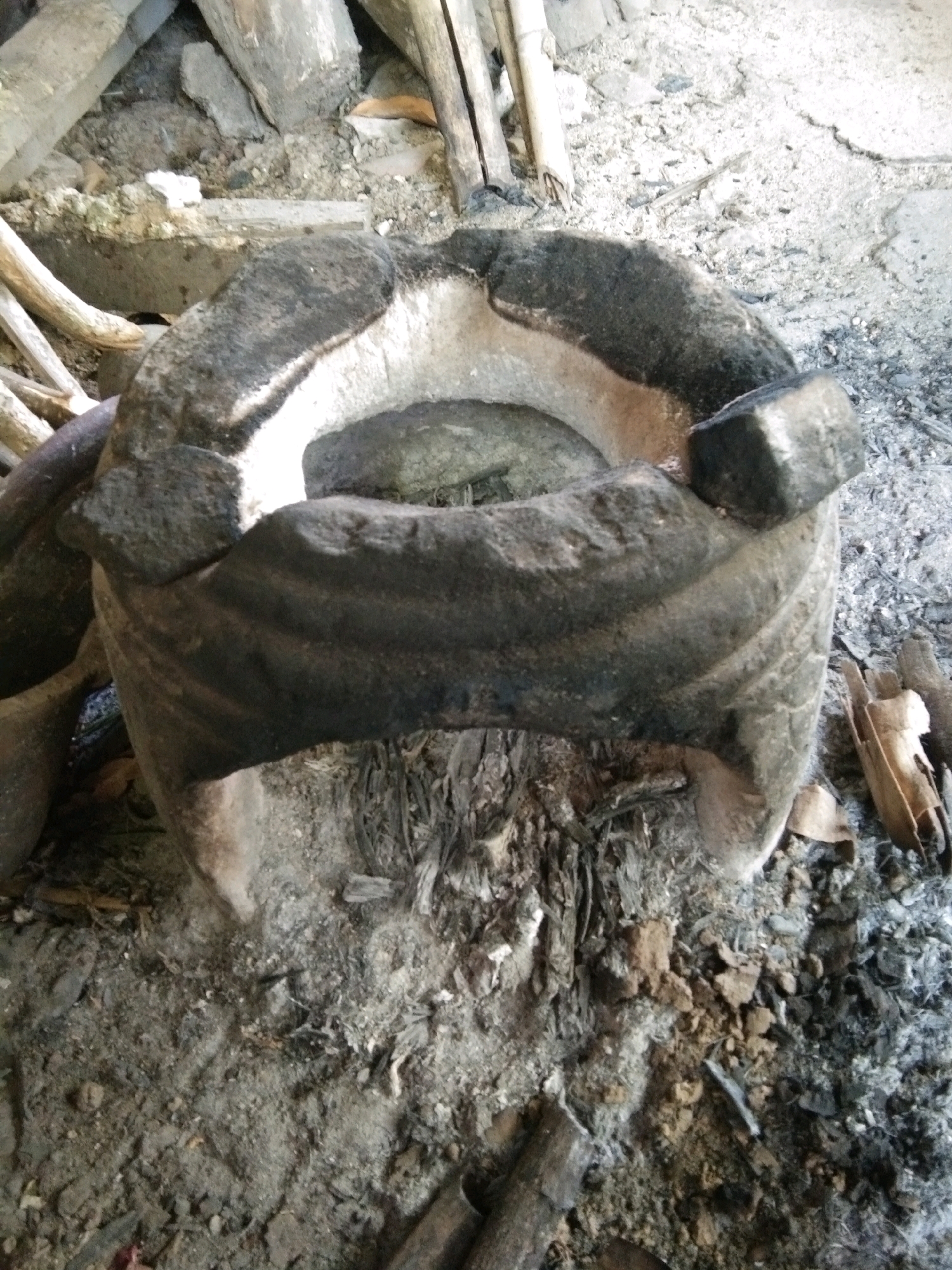 A close-up, overhead shot shows a primitive, soot-covered clay stove used for cooking or smelting, situated in an outdoor, rural setting. The stove is dark gray and weathered, with a wide opening at the top revealing a lighter-colored interior lining. It rests on a bed of ash and debris, surrounded by dry leaves, twigs, and what appear to be parts of broken pottery or bricks. In the background, pieces of wood and bamboo are visible, hinting at a rustic or makeshift environment. The lighting suggests it is daytime, with natural light illuminating the scene. The overall impression is one of an old, well-used cooking or artisanal setup in a humble dwelling or workshop, likely in the rural area of Karangsambung, Indonesia, given the context. There are no people or animals visible in the frame.