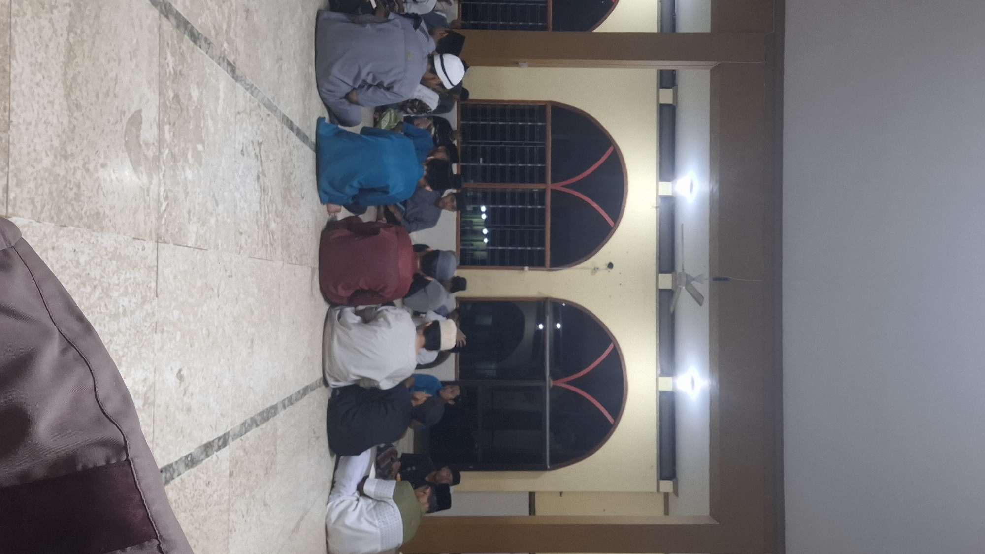 A group of at least 15 males, appearing to be children and young adults, are sitting on a light-colored tiled floor in a semi-circle. They are facing away from the camera, towards a wall with arched windows that are covered by metal grates and curtains. The setting appears to be indoors, possibly a mosque or a religious school in Wonorejo, Indonesia, given the architectural style of the windows and the attire of the individuals. The males are dressed in traditional Islamic clothing, including peci caps and kurtas in various colors such as white, blue, gray, and maroon. Some are wearing white skullcaps, while others have a white kopyah or a white turban-like head covering. The lighting in the room is a mix of overhead ceiling lights and what appear to be recessed lights. A ceiling fan is visible in the upper portion of the frame. The floor is tiled with a marble-like pattern. The background shows the continuation of the wall and the arched windows. The overall atmosphere suggests a gathering for prayer, study, or a religious event.