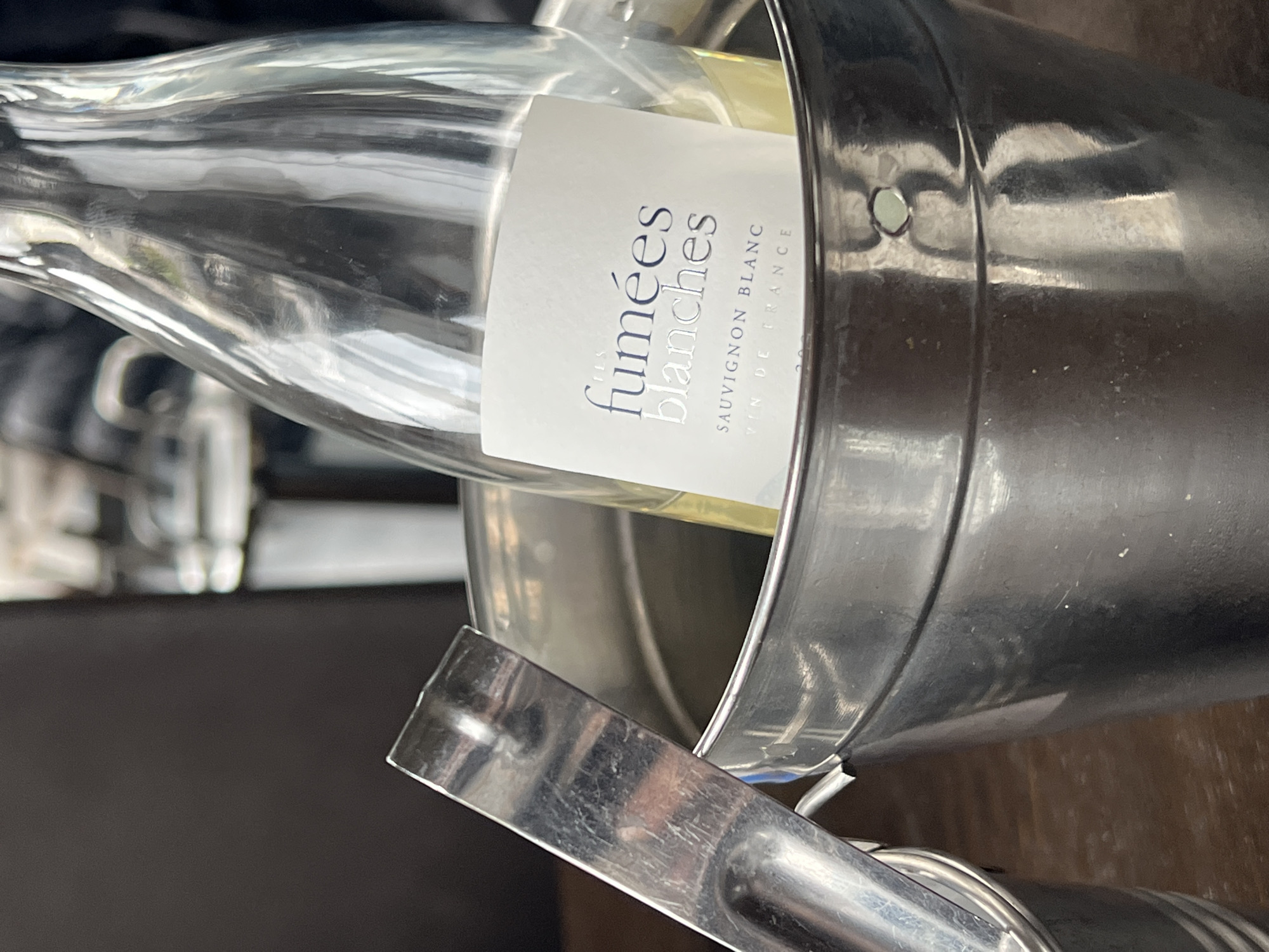 A bottle of "Fumees Blanches" Sauvignon Blanc wine is situated in a silver ice bucket, possibly at a restaurant or bar. The image is captured at an oblique angle, with the bottle's label visible. The scene is blurry in the background. No specific details suggest the location is Sofia, Bulgaria.