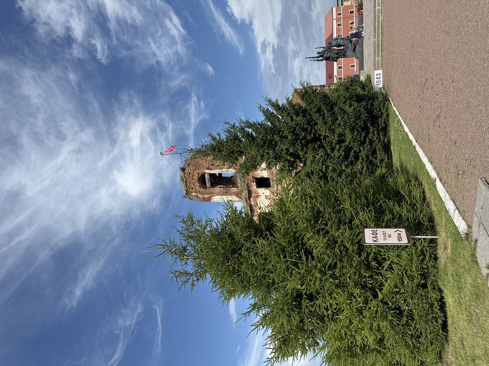 The image captures a view of the Shlisselburg Fortress, also known as Oreshek Fortress, in Shlisselburg, Russia. In the foreground, a large, vibrant green spruce tree dominates the left side of the frame, with another similar tree partially obscuring the background. A white sign with maroon trim is visible to the right of the spruce, bearing Russian text that translates to "CAFE TOILET WC 150m" with an arrow pointing right.
Behind the trees, the weathered brick ruins of a fortress structure are prominent, showing signs of decay and damage. A bright red flag flies from a pole atop the ruins. To the right of the ruins, a monument featuring several bronze figures holding rifles stands on a raised platform, with the year "1943" inscribed on a stone block in front of it. In the far background, several red-roofed buildings with white window frames are visible. The sky is a bright blue, streaked with thin, wispy white clouds. The ground in the foreground is a mixture of gravel and paved areas. Two people, a child and an adult, are partially visible near the monument in the mid-ground.