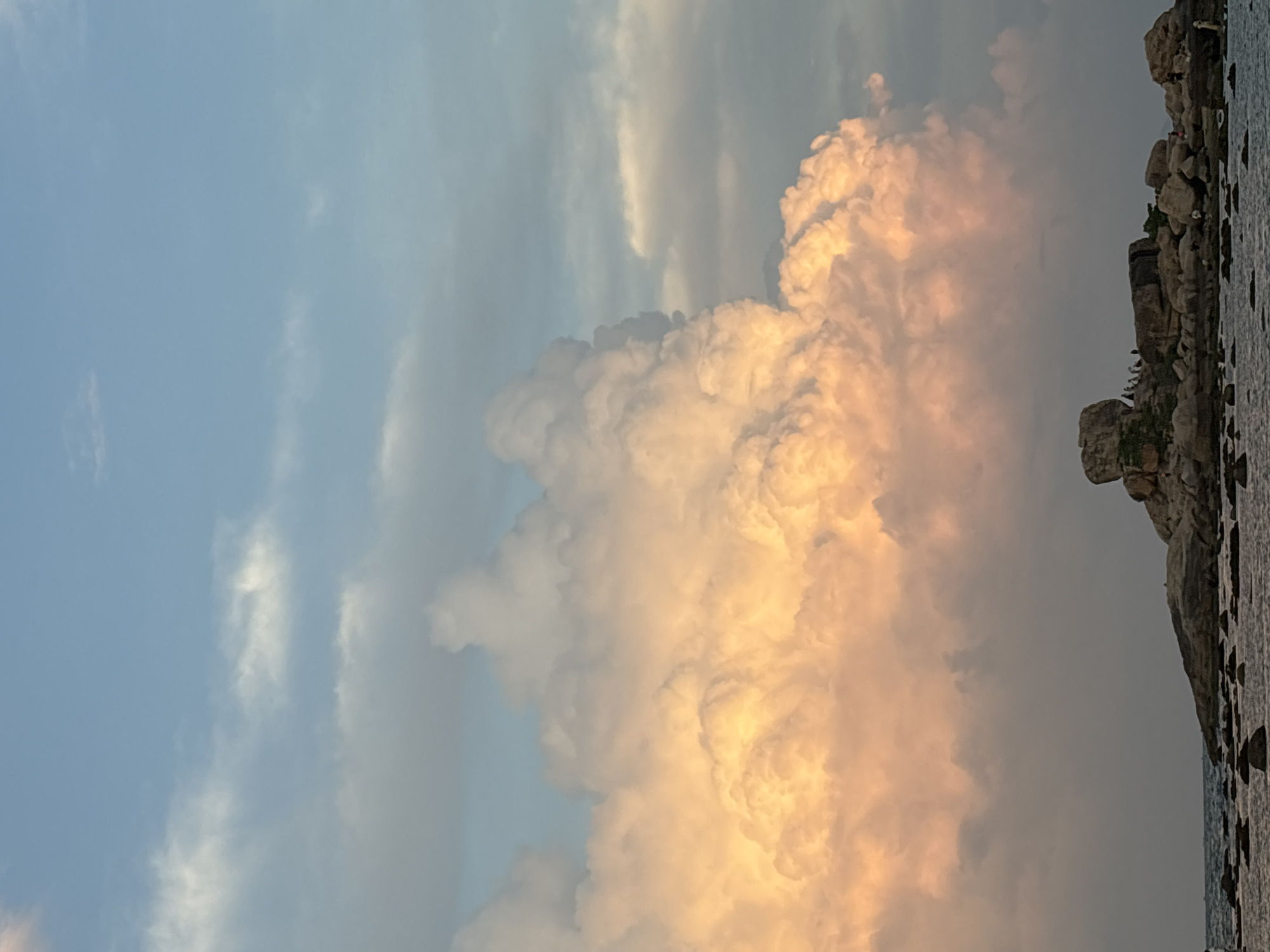 Captured in Bac Nha Trang Ward, Vietnam, the image portrays a sky dominated by a large, billowing cumulus cloud formation. The cloud is illuminated with a warm, golden light, likely from the setting sun. Adjacent to the cloud, on the right side of the image, is a rocky shoreline with visible water.